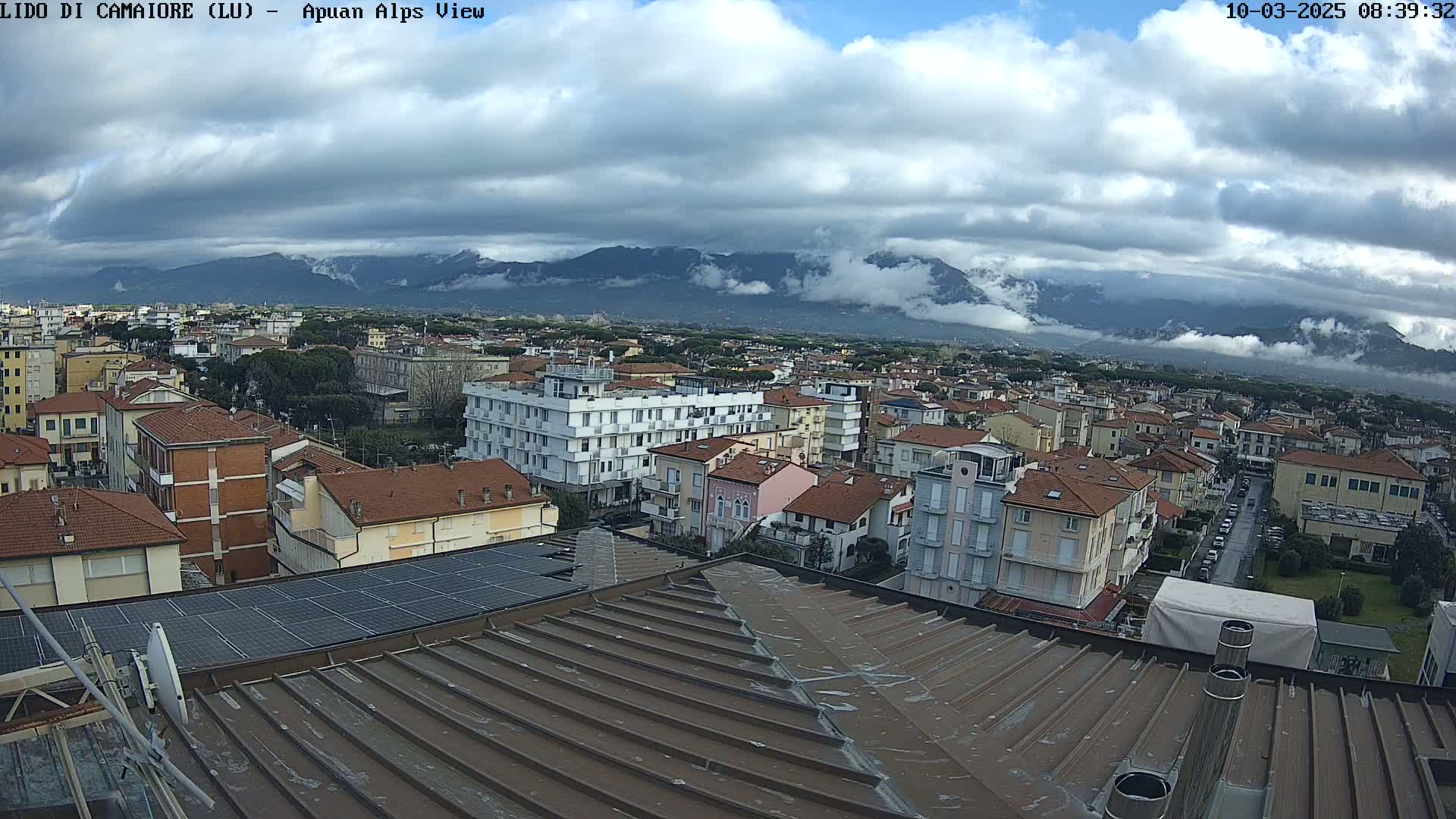 A view from a rooftop shows a town of low-rise buildings under a cloudy sky, with mountains partially obscured by fog in the distance.