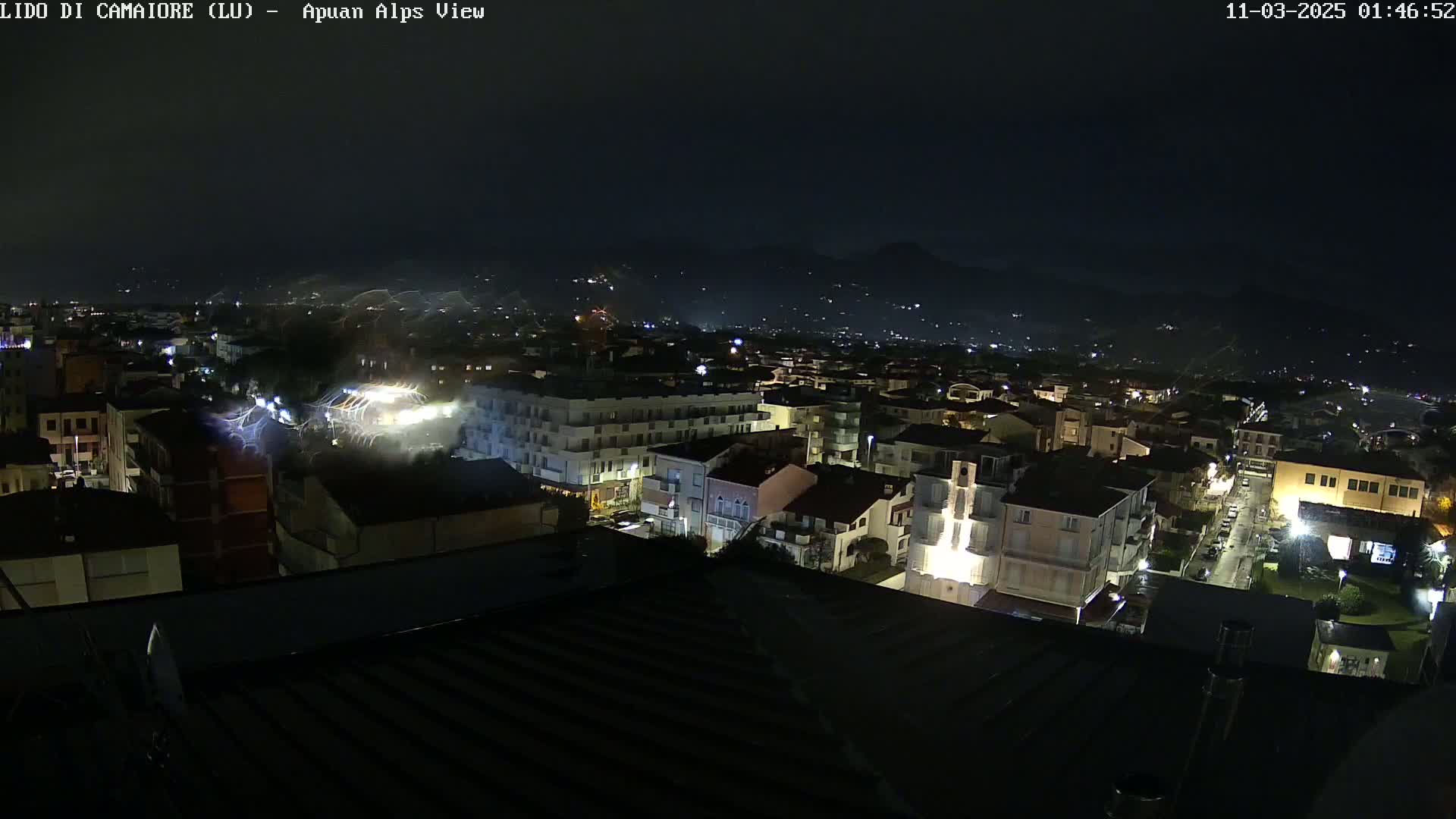 A nighttime aerial view of a town, illuminated by streetlights, with faint streaks of light possibly from rain or other atmospheric phenomena, and dark mountains visible in the background.