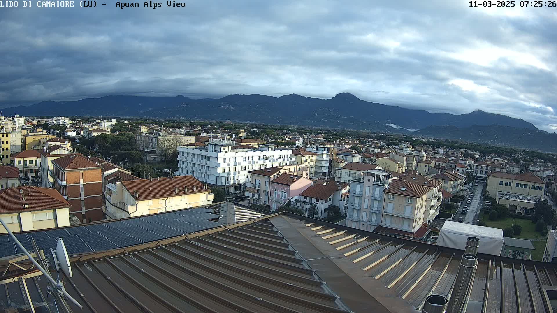 A view from a rooftop shows a town nestled at the foot of mountains under a cloudy sky.
