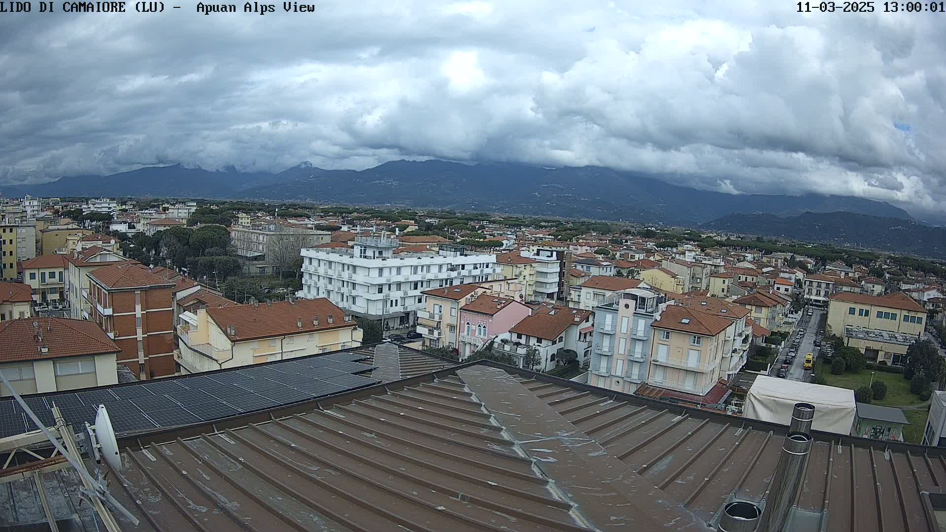 A cloudy day reveals a cityscape of low-rise buildings, a distant mountain range, and a rooftop view with solar panels.