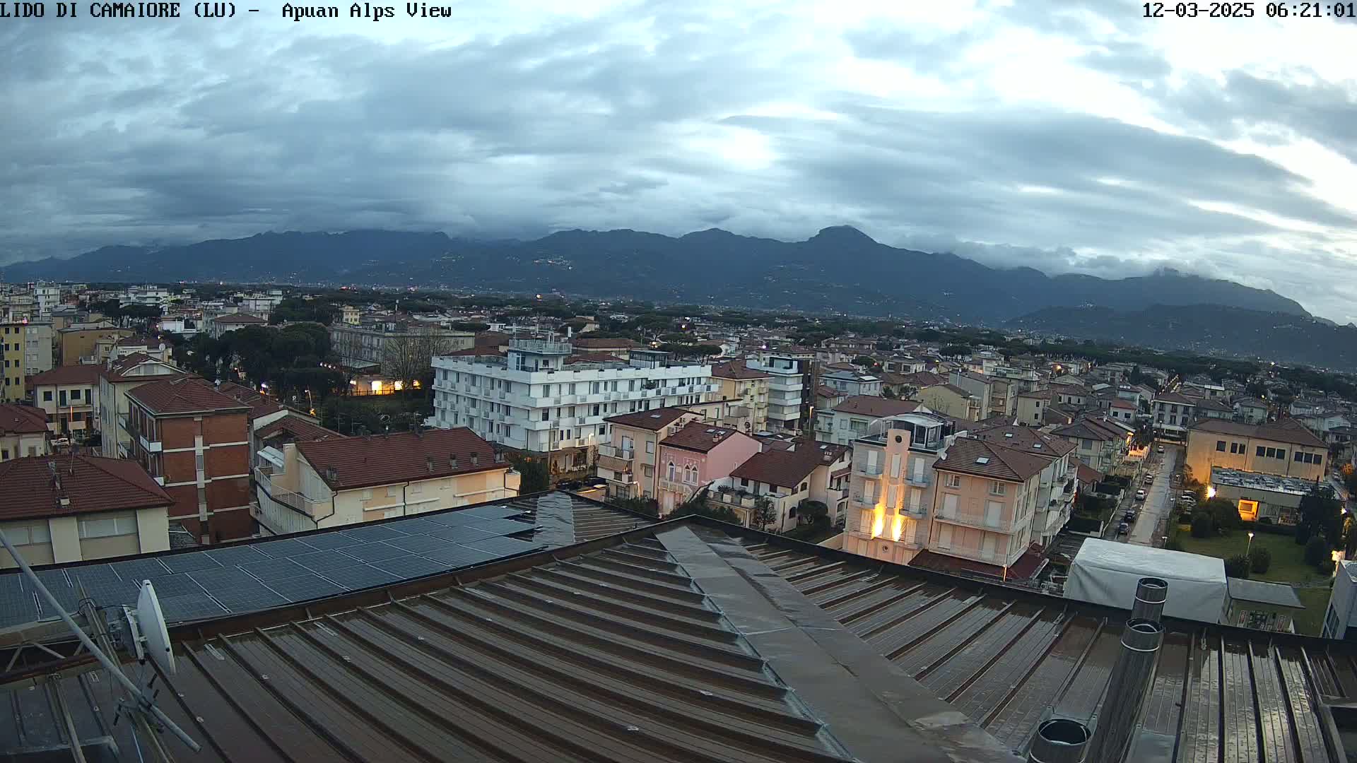 A wet rooftop overlooks a town nestled beneath a mountain range under a cloudy sky.