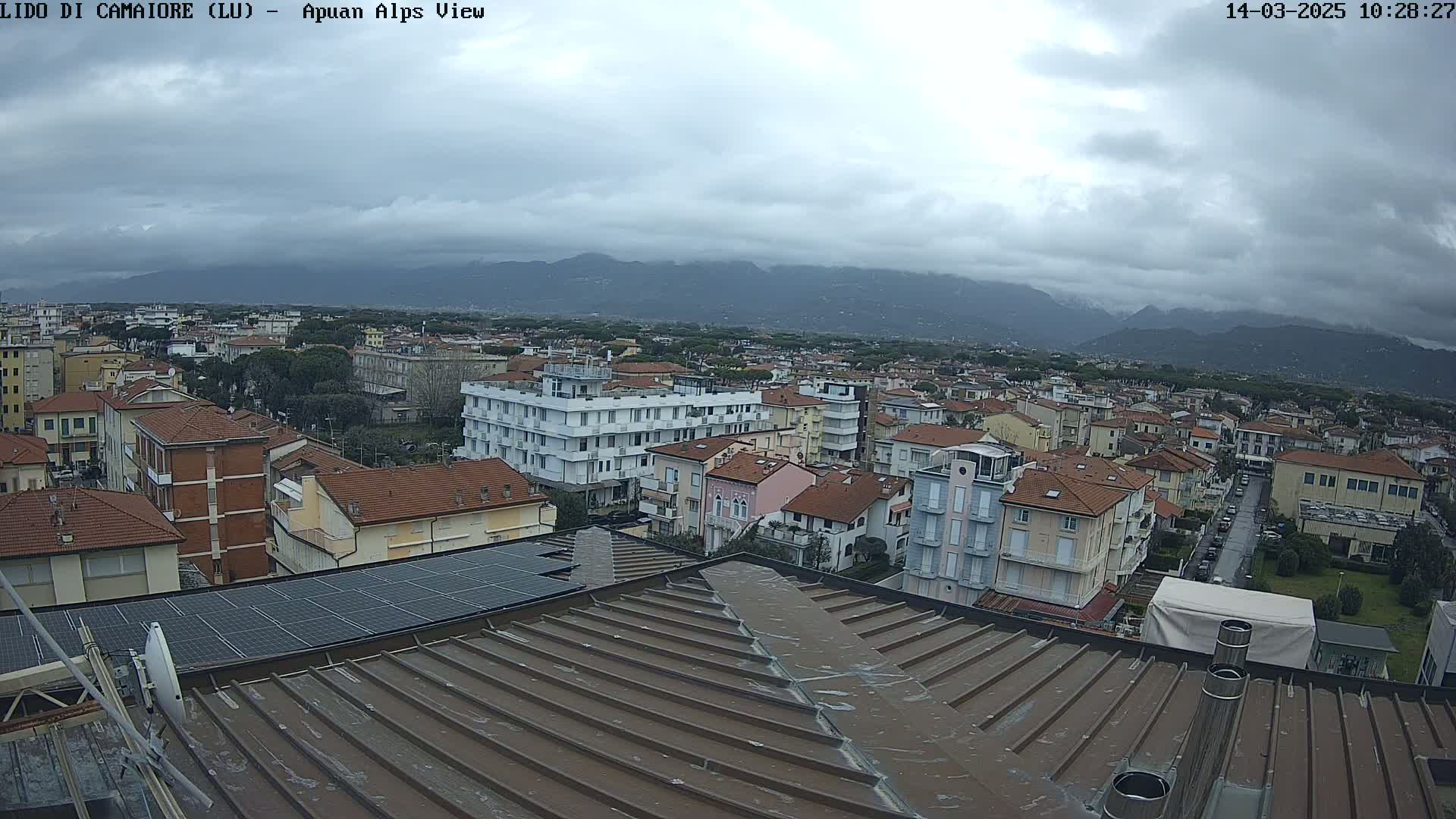 A view from a rooftop shows a town under a cloudy sky, with mountains in the background.