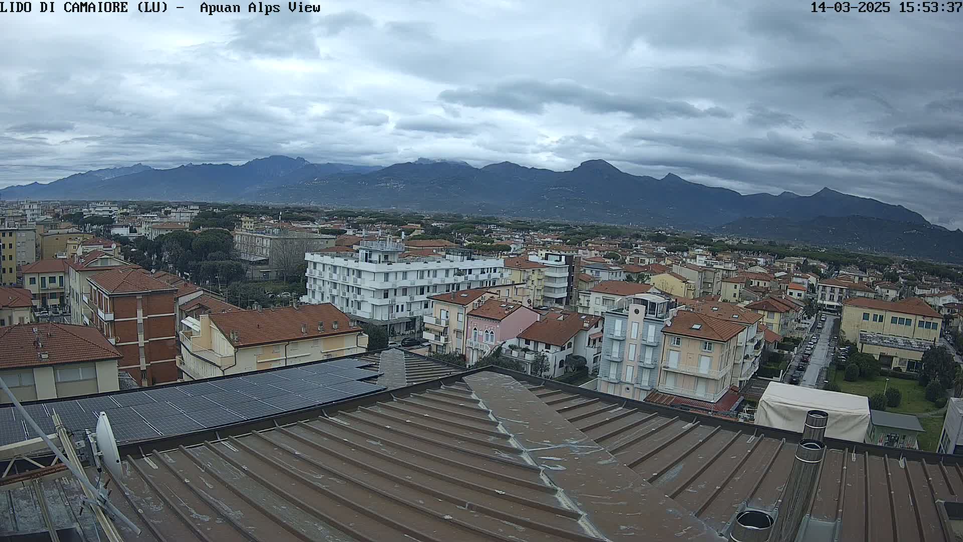 A wide shot from a rooftop shows a town with low-rise buildings under a cloudy sky, with mountains visible in the distance.