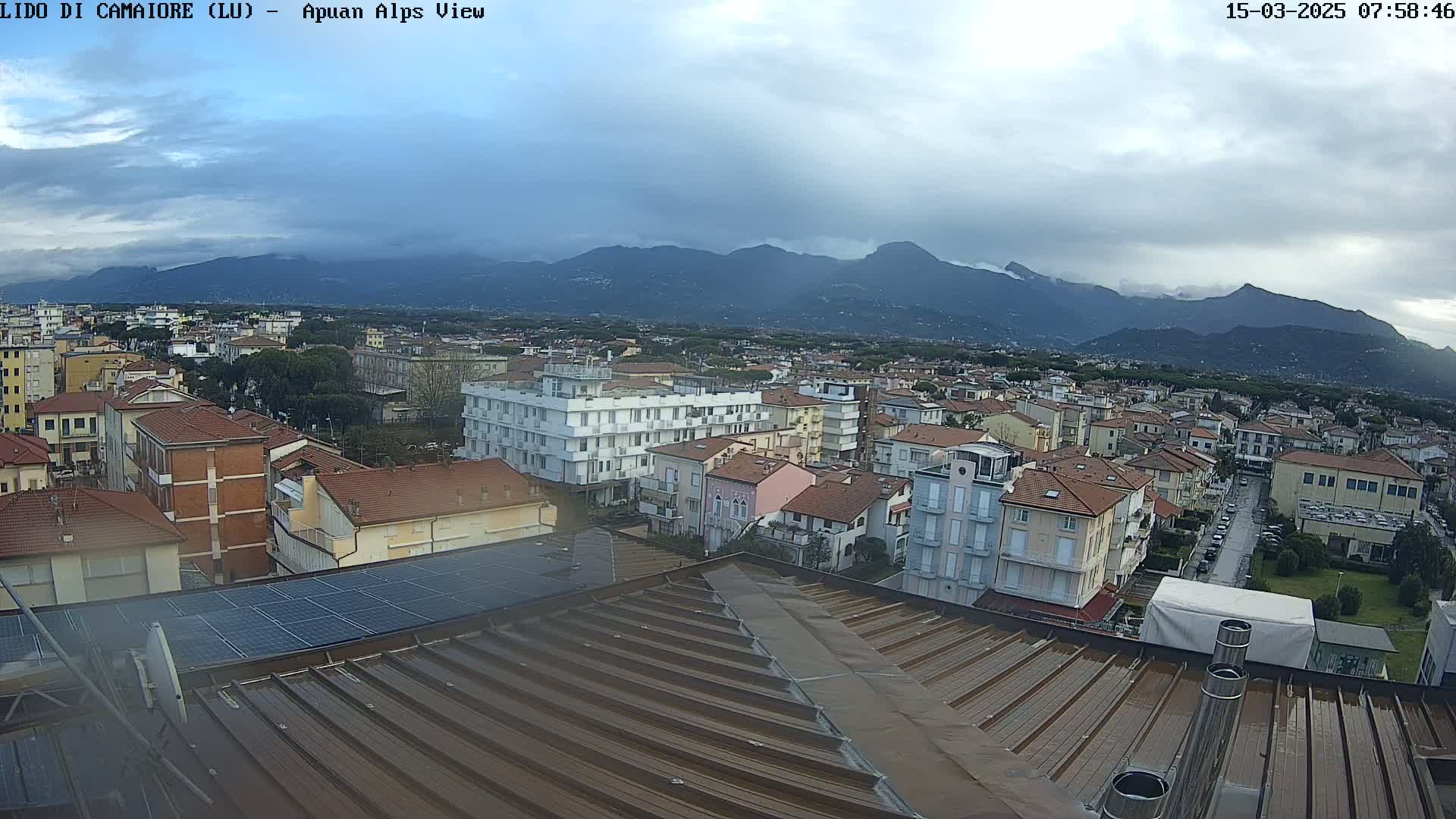 A view of a town with red-tiled roofs and a mountain range in the background under an overcast sky.