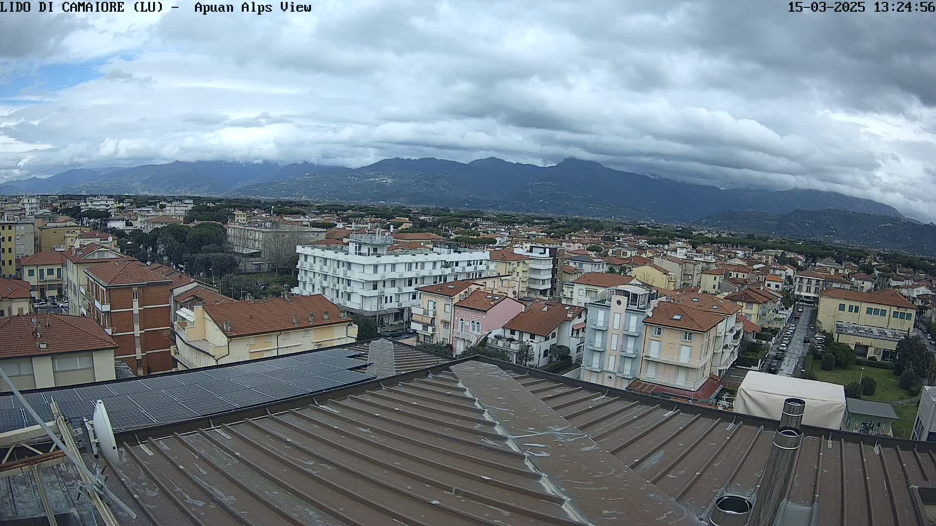 A view from a rooftop shows a town nestled beneath a cloudy sky and a mountain range in the distance.