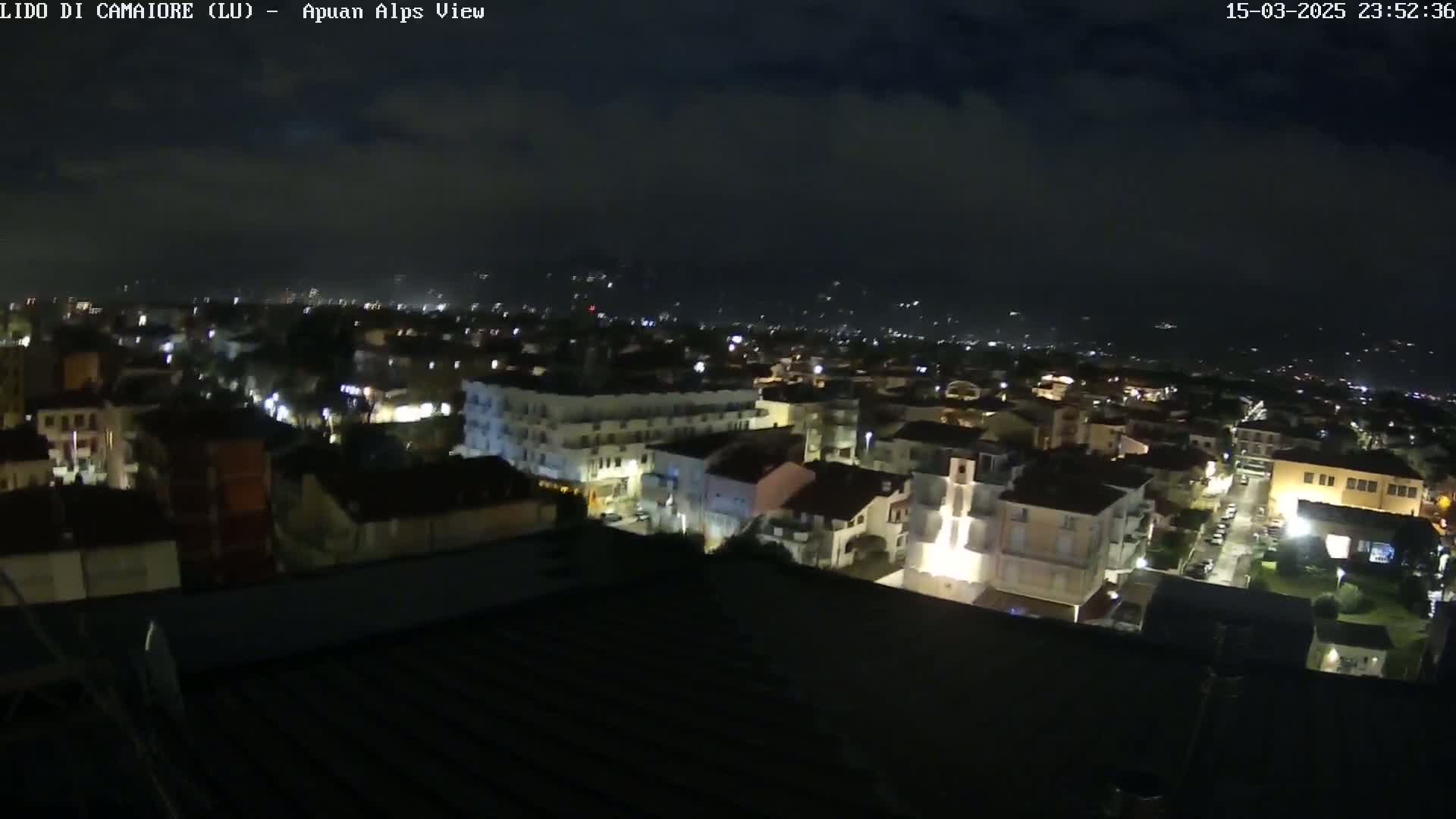 A nighttime aerial view of a town shows many buildings with lights on, under a partly cloudy sky.