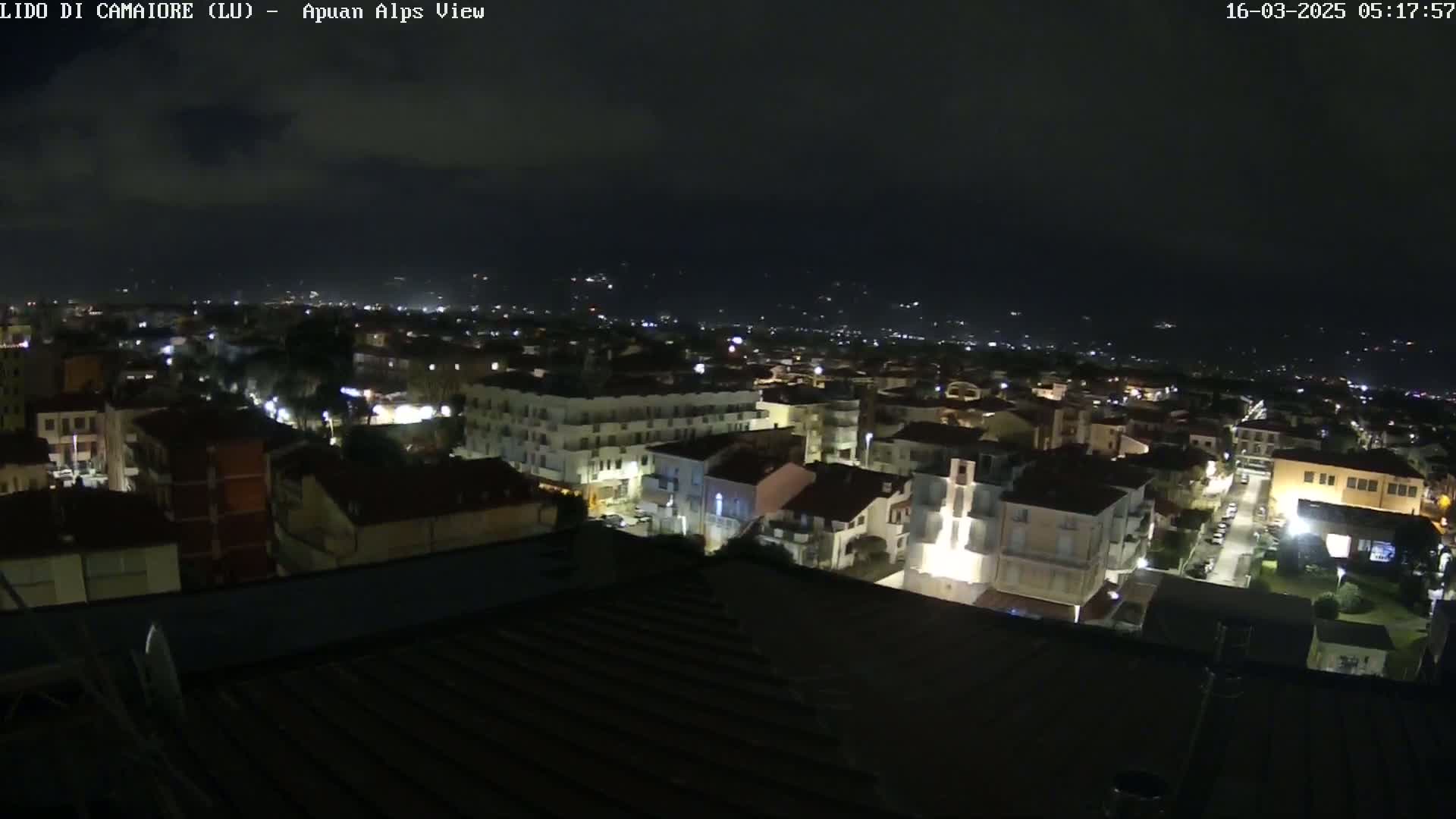 A nighttime aerial view of a town with many illuminated buildings, under a dark, mostly cloudy sky.
