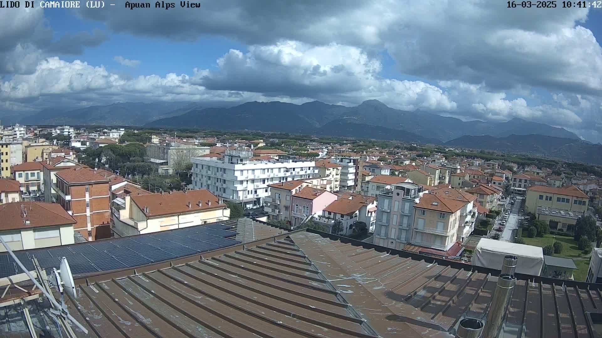 A partly cloudy day reveals a sprawling town of low-rise buildings nestled at the foot of a mountain range, viewed from a rooftop with solar panels.