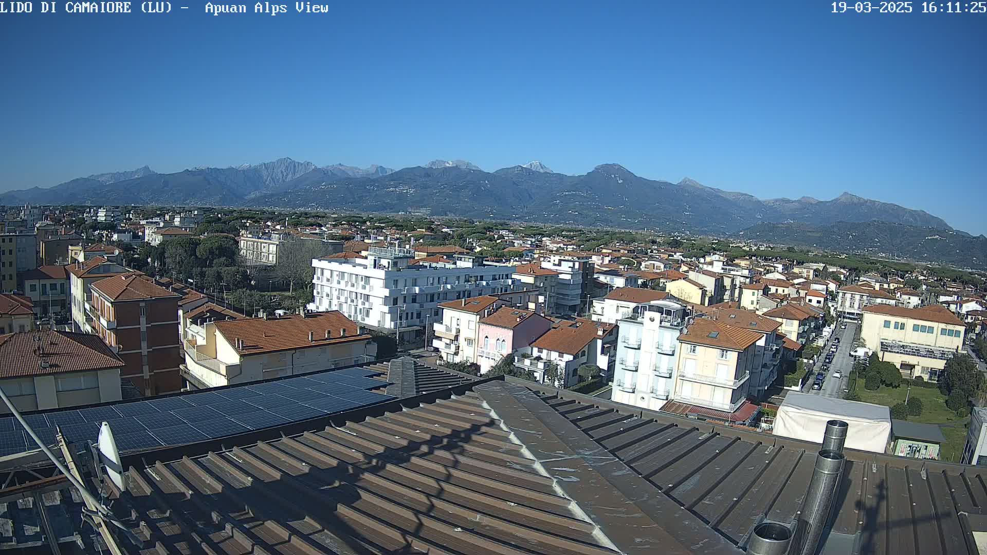 A clear, sunny day reveals a view of a coastal town nestled at the foot of a mountain range, seen from a rooftop with solar panels.