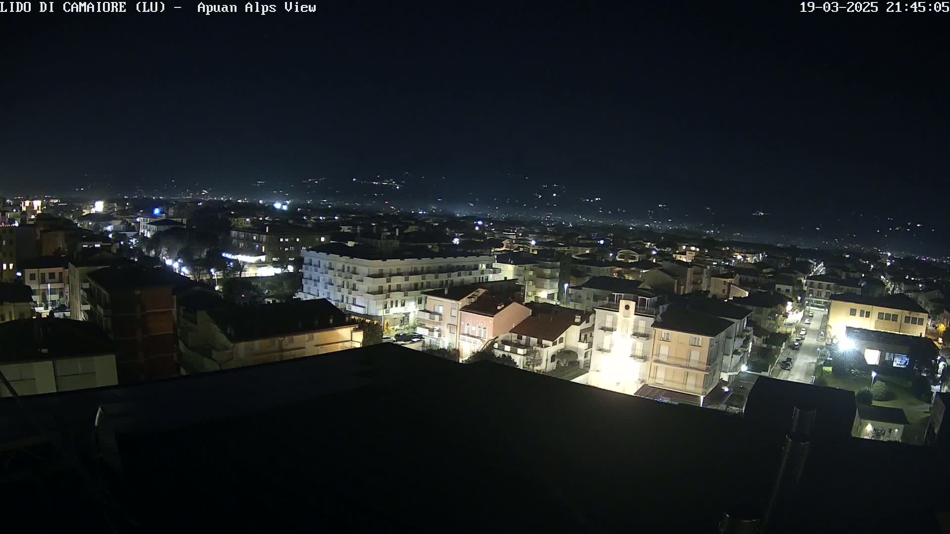 A nighttime aerial view of a town, illuminated by streetlights, with a distant mountain range visible under a clear, dark sky.