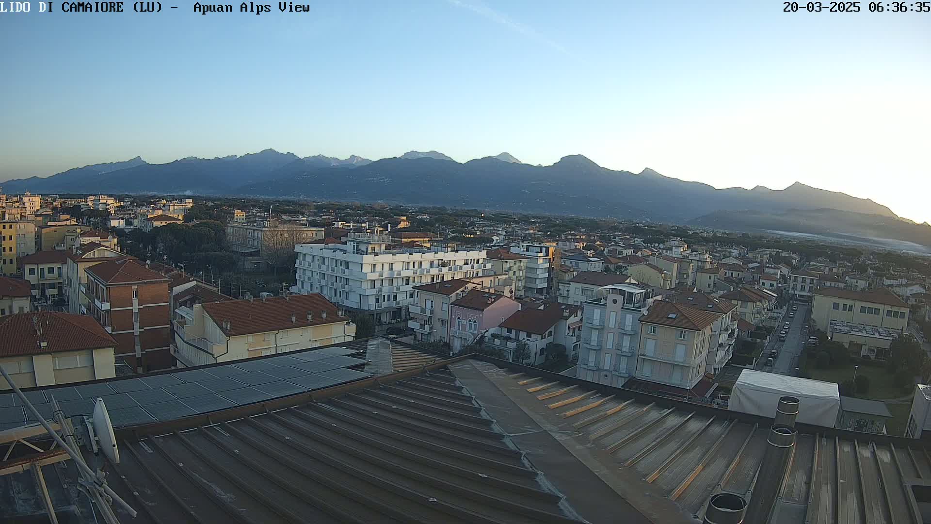 A clear sunrise illuminates a town nestled below a mountain range, viewed from a rooftop with solar panels.