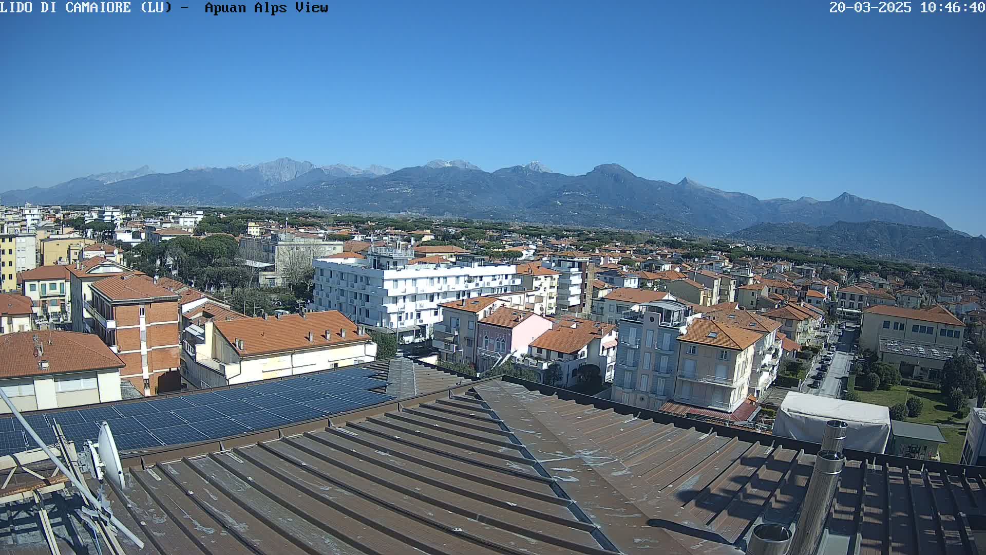 A clear, sunny day reveals a cityscape nestled at the foot of a mountain range, viewed from a rooftop with solar panels.