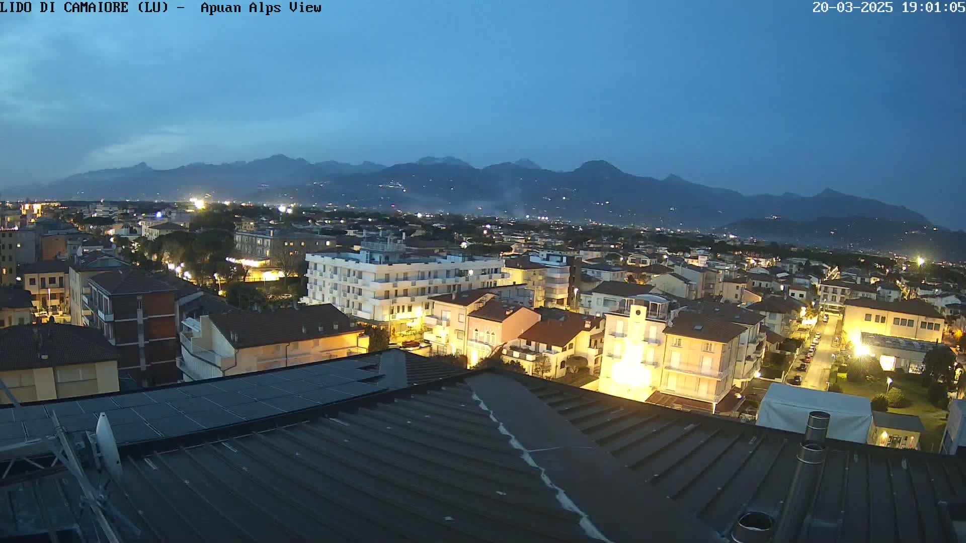 A nighttime view of a town illuminated by streetlights, with a mountain range visible in the background under a twilight sky.