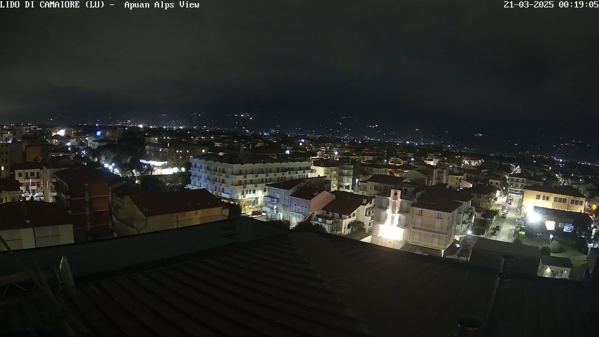 A nighttime aerial view of a town with illuminated buildings, streets, and distant hills under a dark, clear sky.