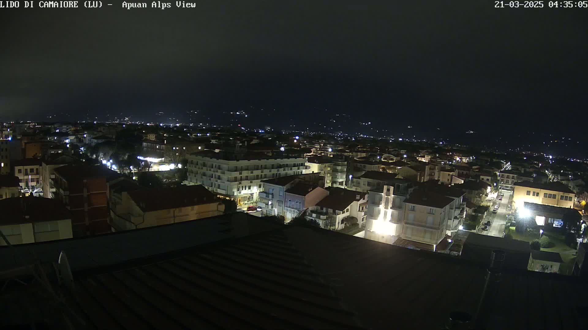 A nighttime aerial view of a town illuminated by streetlights, with a dark sky and distant hills faintly visible in the background.