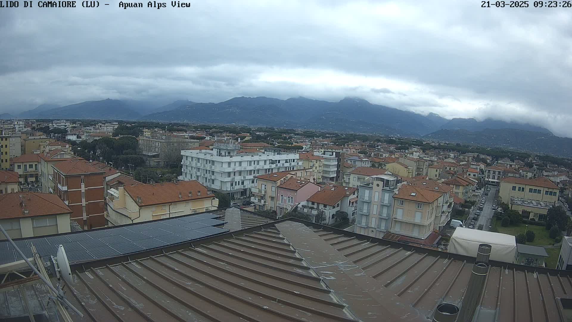 A cloudy day reveals a view of a town nestled at the base of mountains, with rooftops, buildings, and solar panels visible from an elevated vantage point.