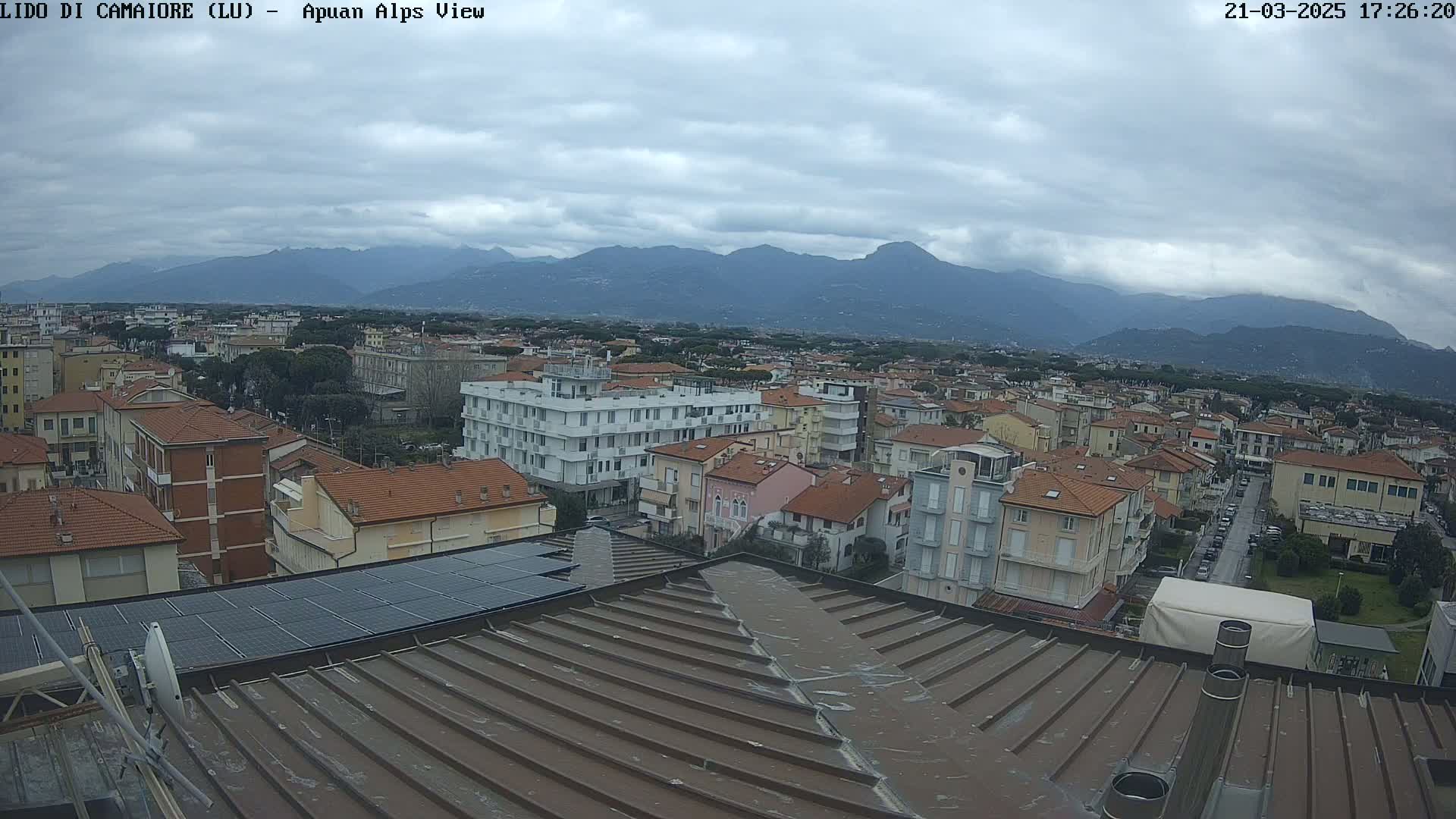 A view from a rooftop shows a town under a cloudy sky with mountains in the distance.