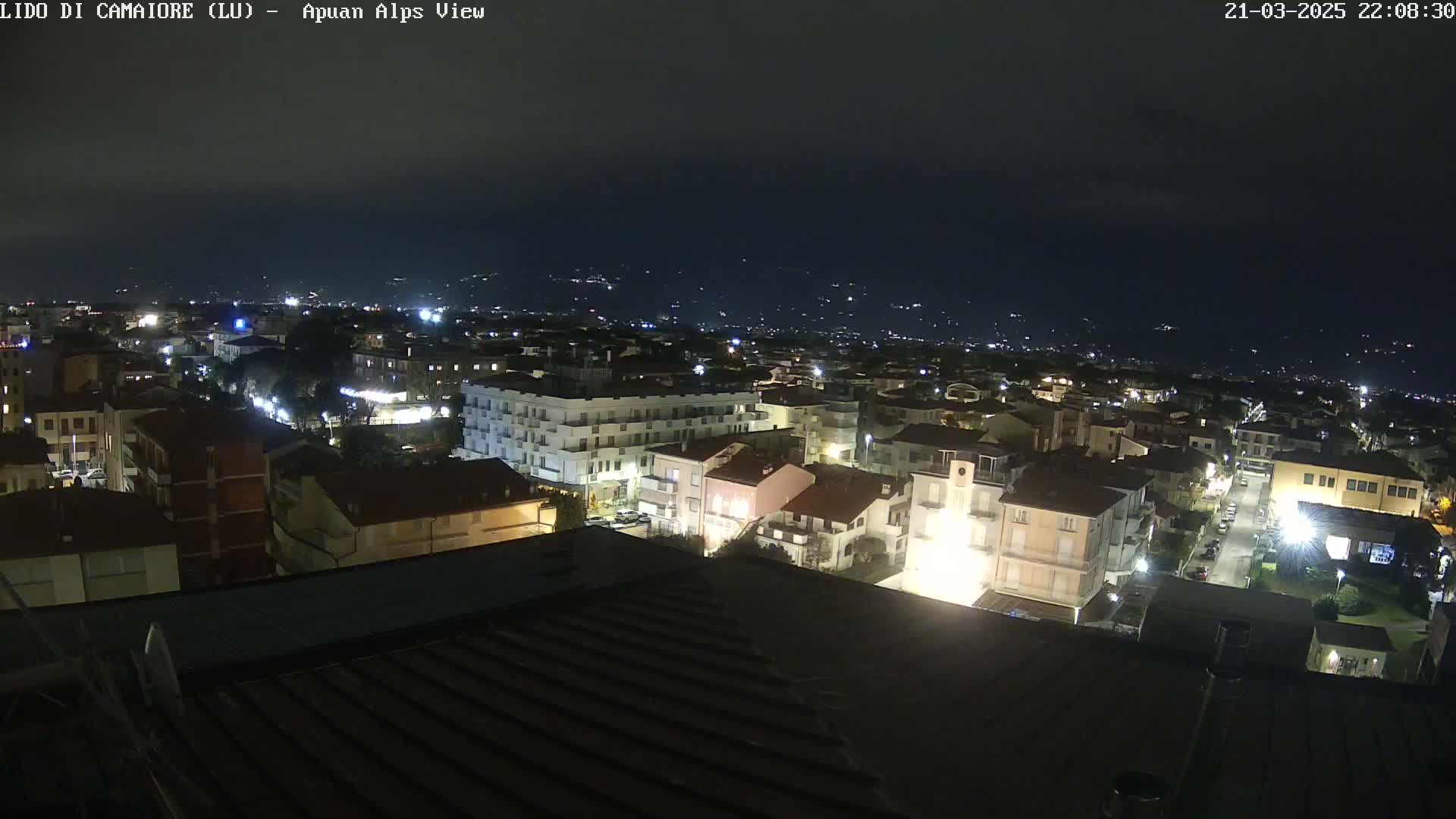 A nighttime, aerial view of a town illuminated by streetlights, with a dark, clear sky and distant hills faintly visible in the background.