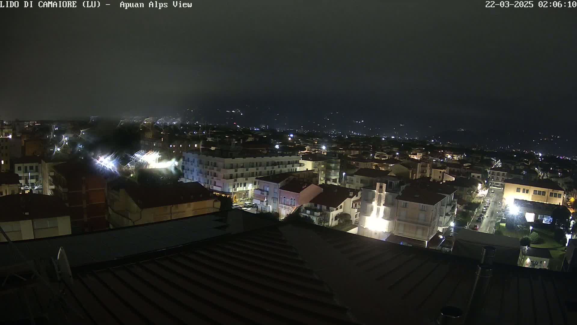A nighttime view of a town illuminated by streetlights, with lightning visible in the distance under a dark, cloudy sky.
