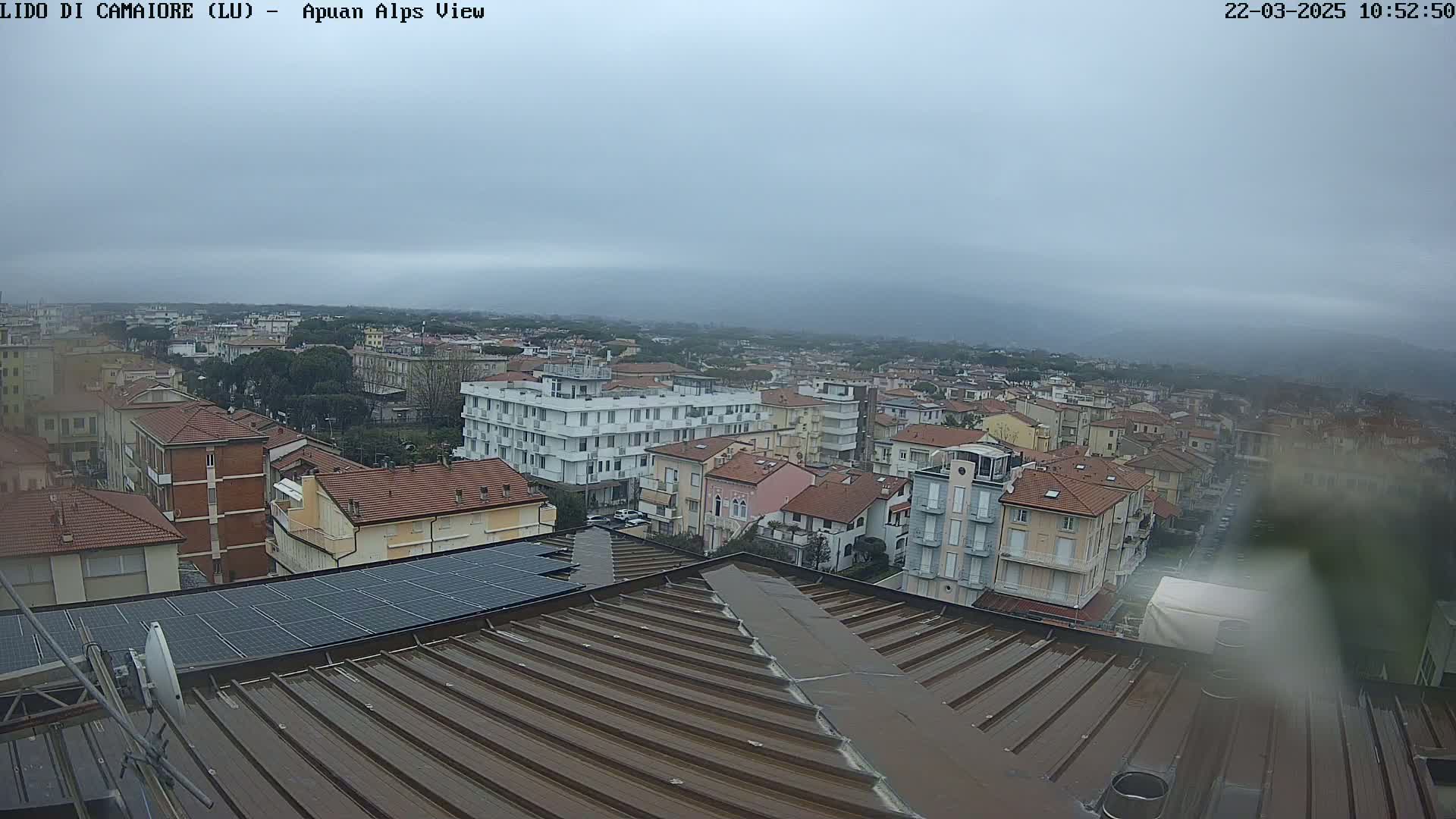 A wet, overcast view of a town, seen from a rooftop with solar panels, showing many multi-story buildings and a distant mountain range.