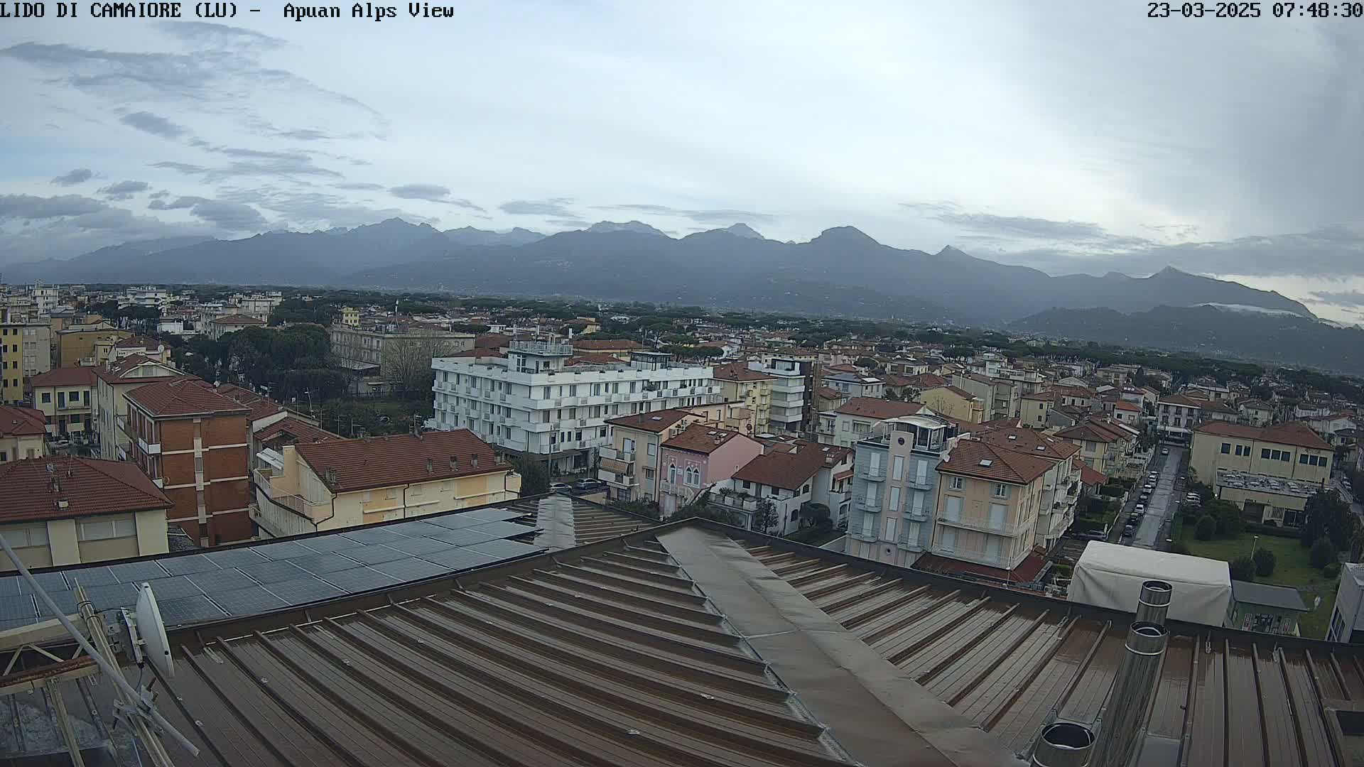 A wet, corrugated metal rooftop overlooks a town nestled against a mountain range under an overcast sky.