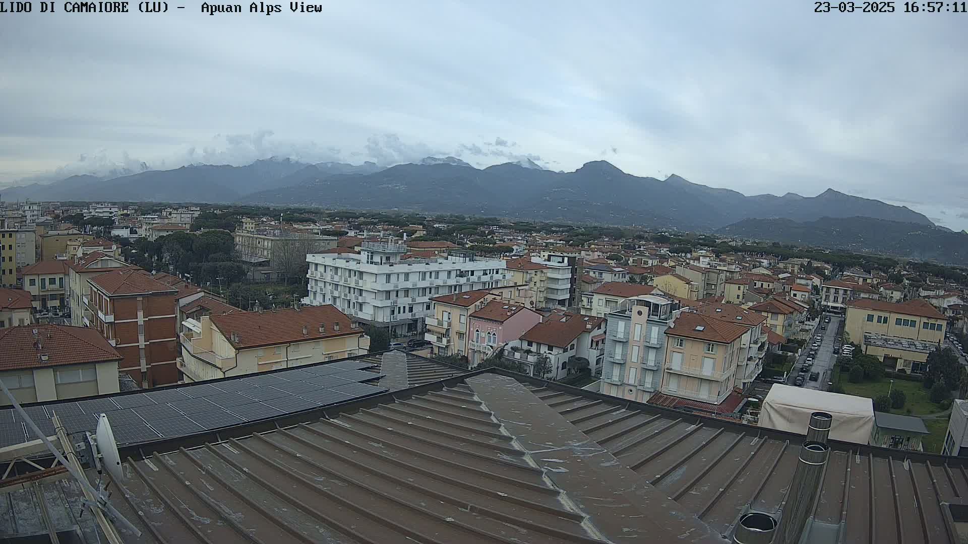 A view from a rooftop shows a town nestled beneath a mountain range on an overcast day.
