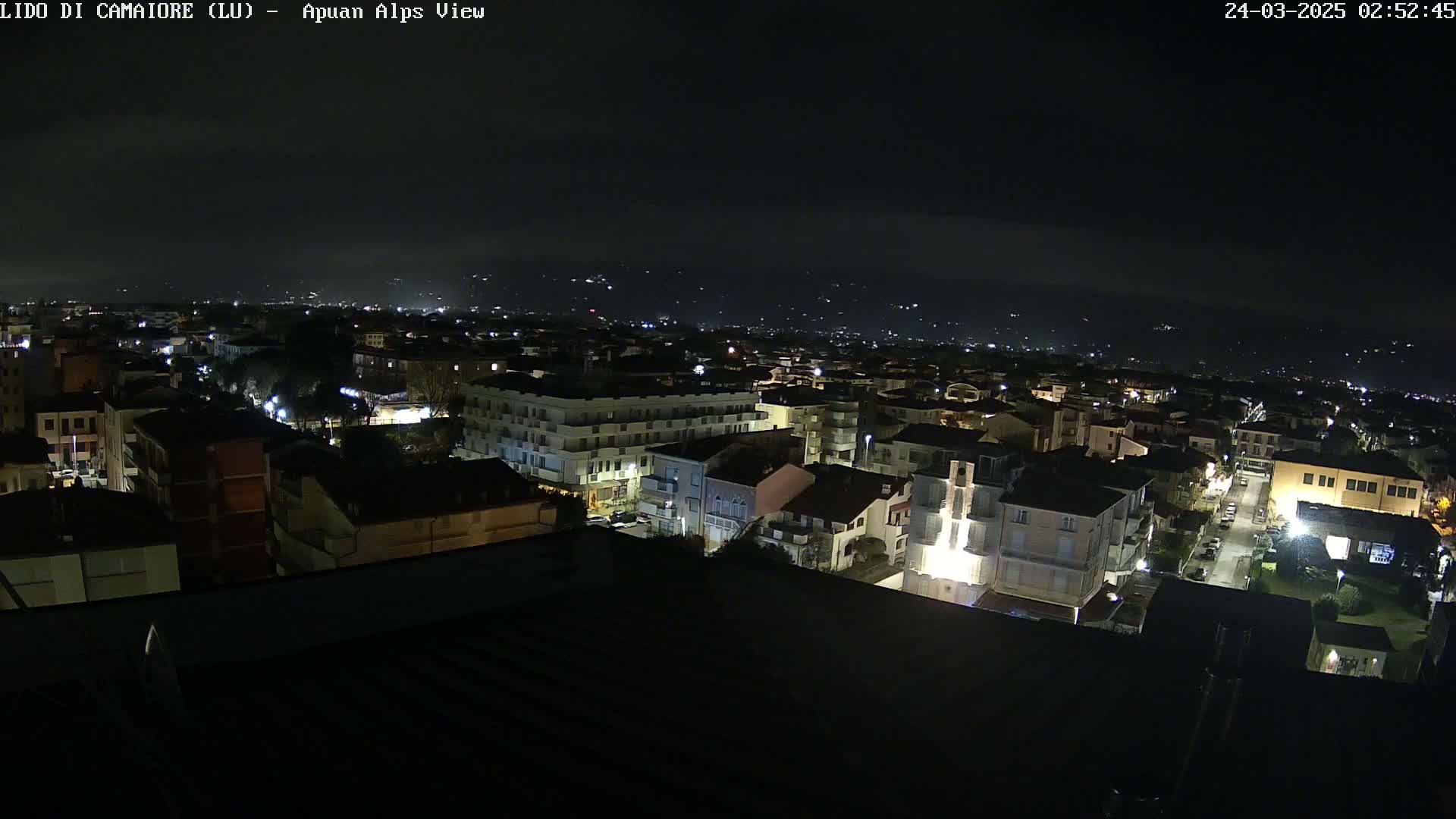 A nighttime aerial view of a town with dimly lit buildings and streets, extending to distant hills under a dark sky.