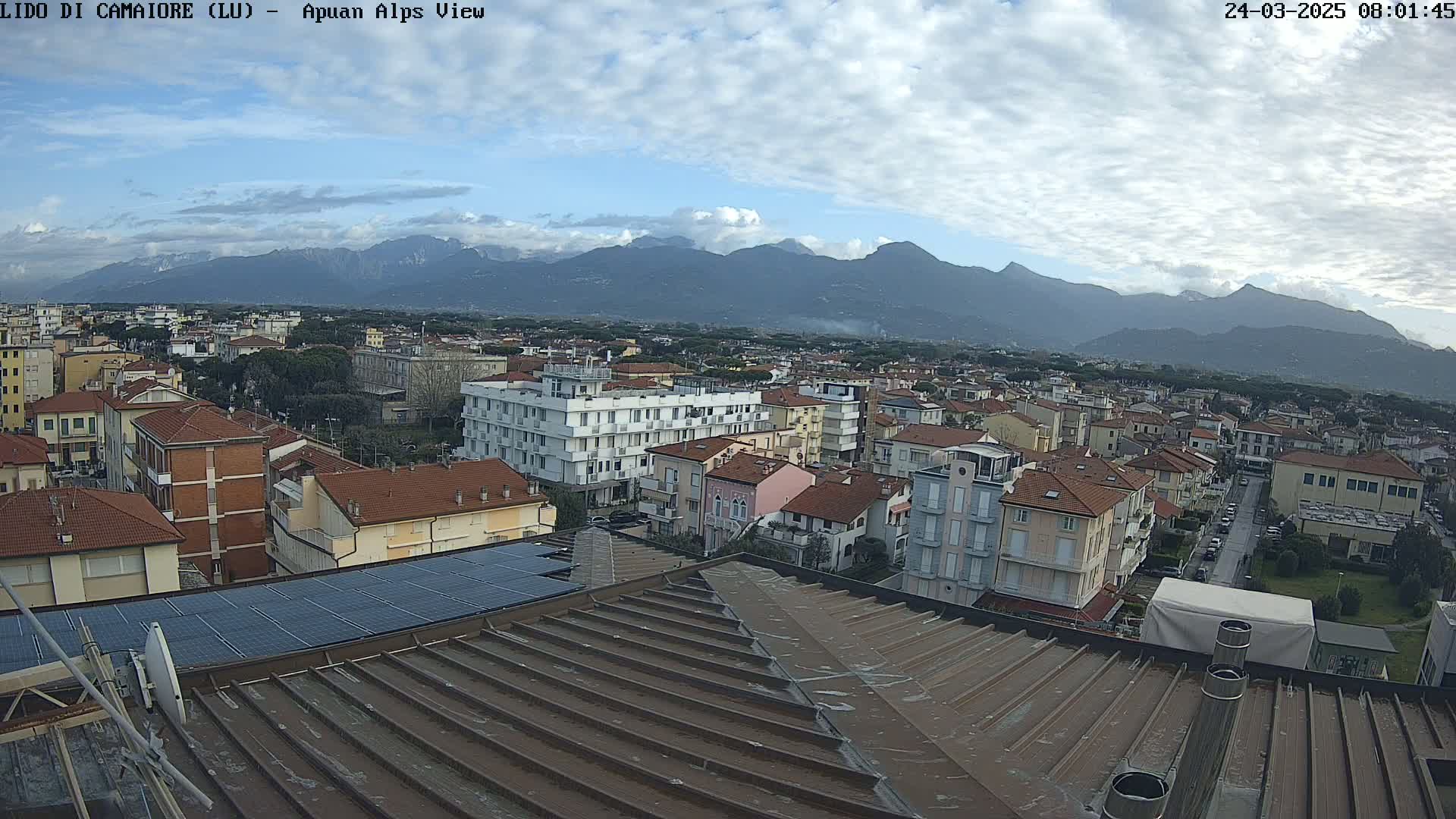 A cloudy day reveals a sprawling town nestled against a mountain range, viewed from a rooftop with solar panels.
