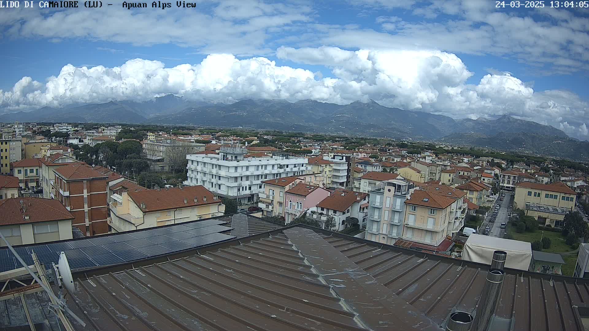 A rooftop view of a town nestled beneath a partly cloudy sky and mountains in the distance.