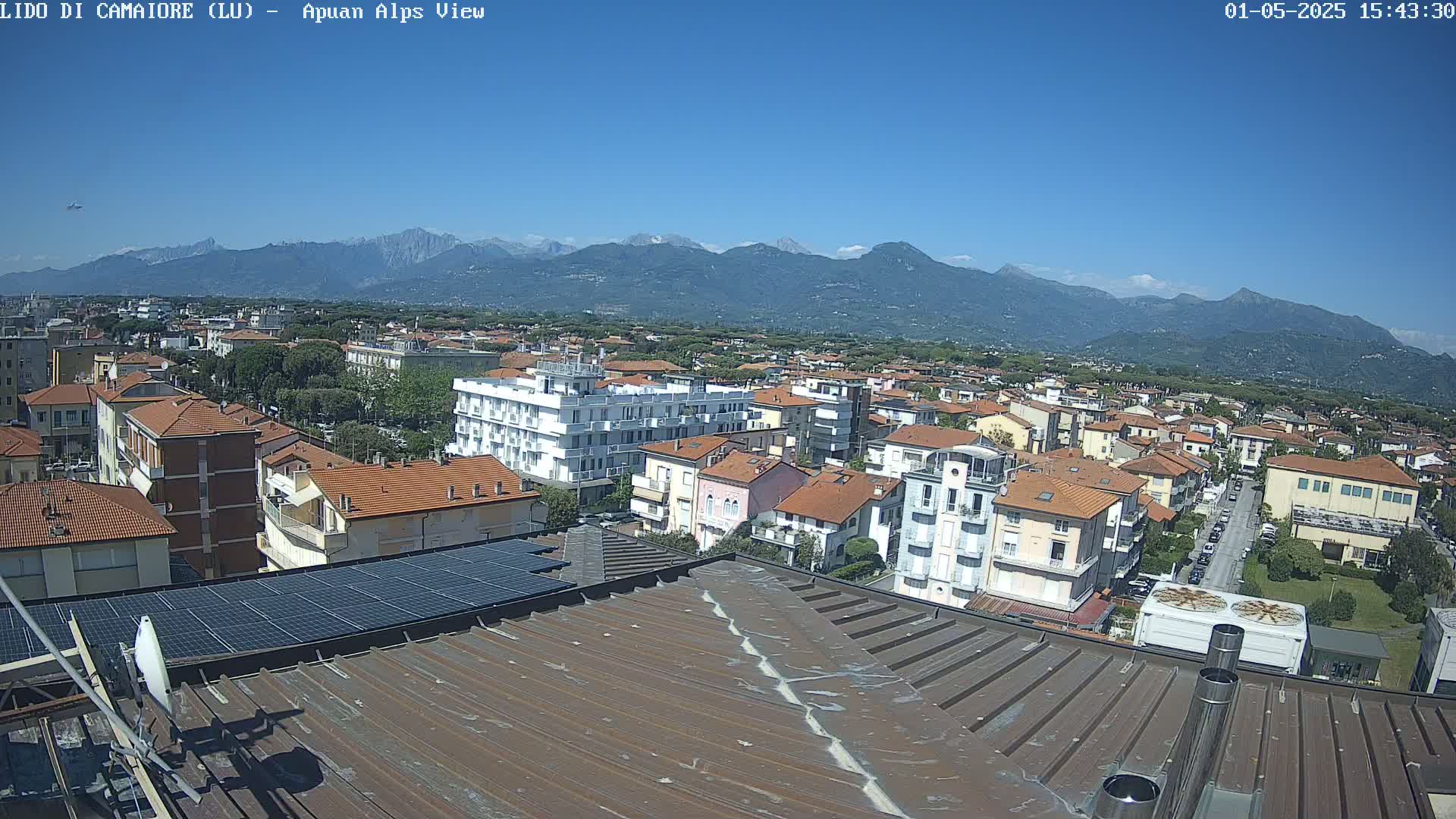 A rooftop view of a town nestled in a valley, overlooked by a mountain range under a clear blue sky.