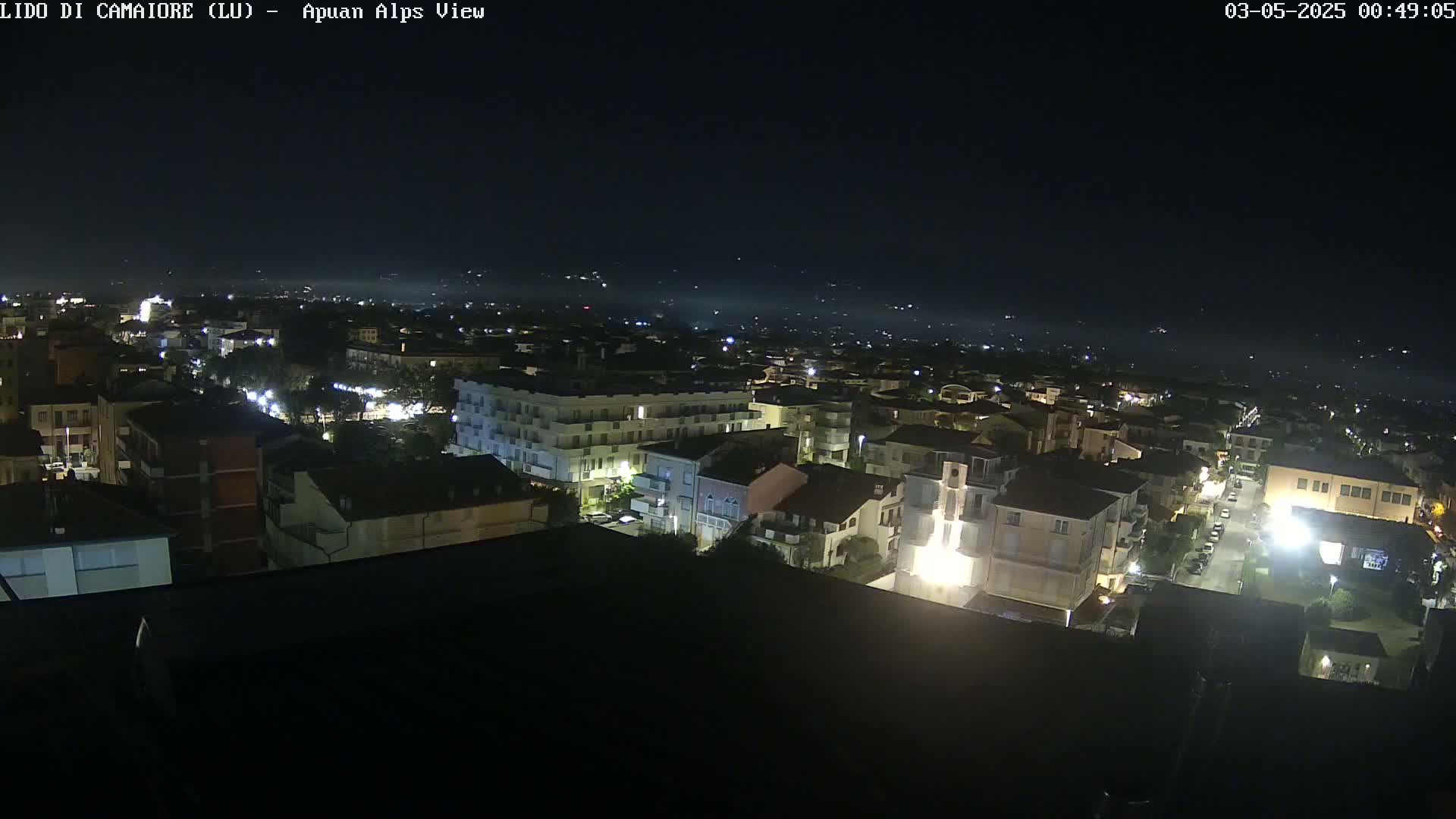 A nighttime aerial view shows a town illuminated with streetlights, extending into a dimly lit landscape under a clear, dark sky.