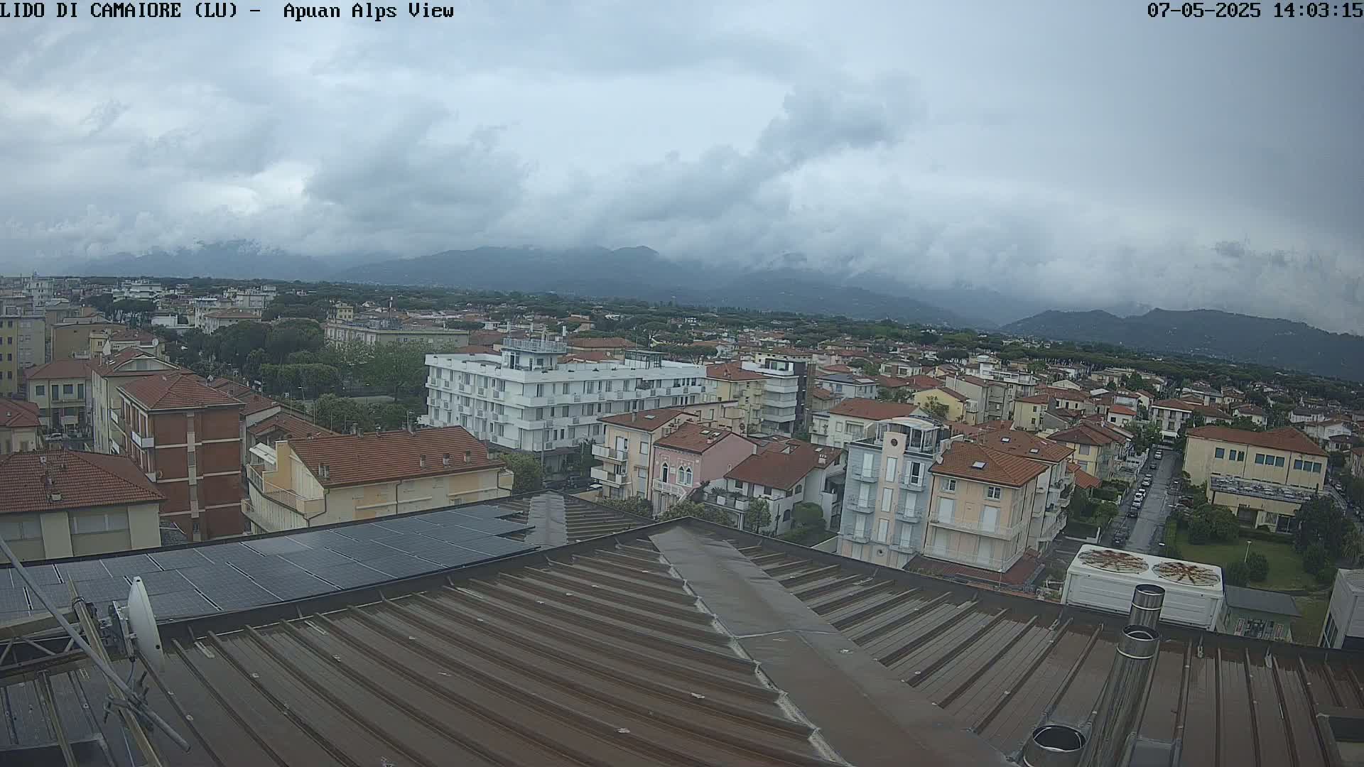 A wide shot of a town under a cloudy sky, showing many buildings and a mountain range in the distance.