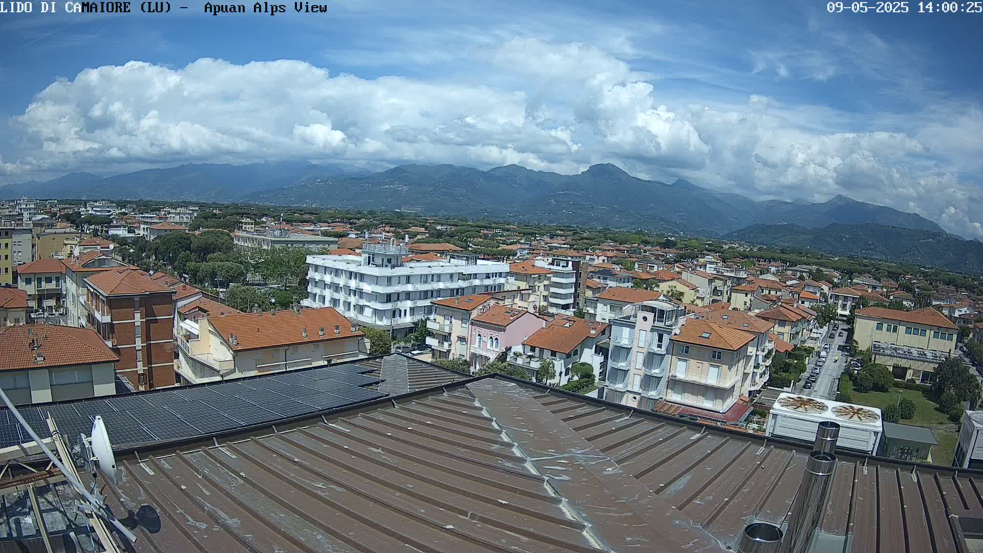 A rooftop view of a town with red-tiled buildings under a partly cloudy sky, showcasing a mountainous landscape in the background.