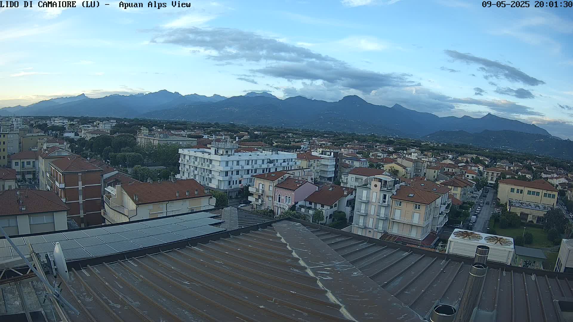 A rooftop view of a town nestled at the base of a mountain range under a partly cloudy evening sky.