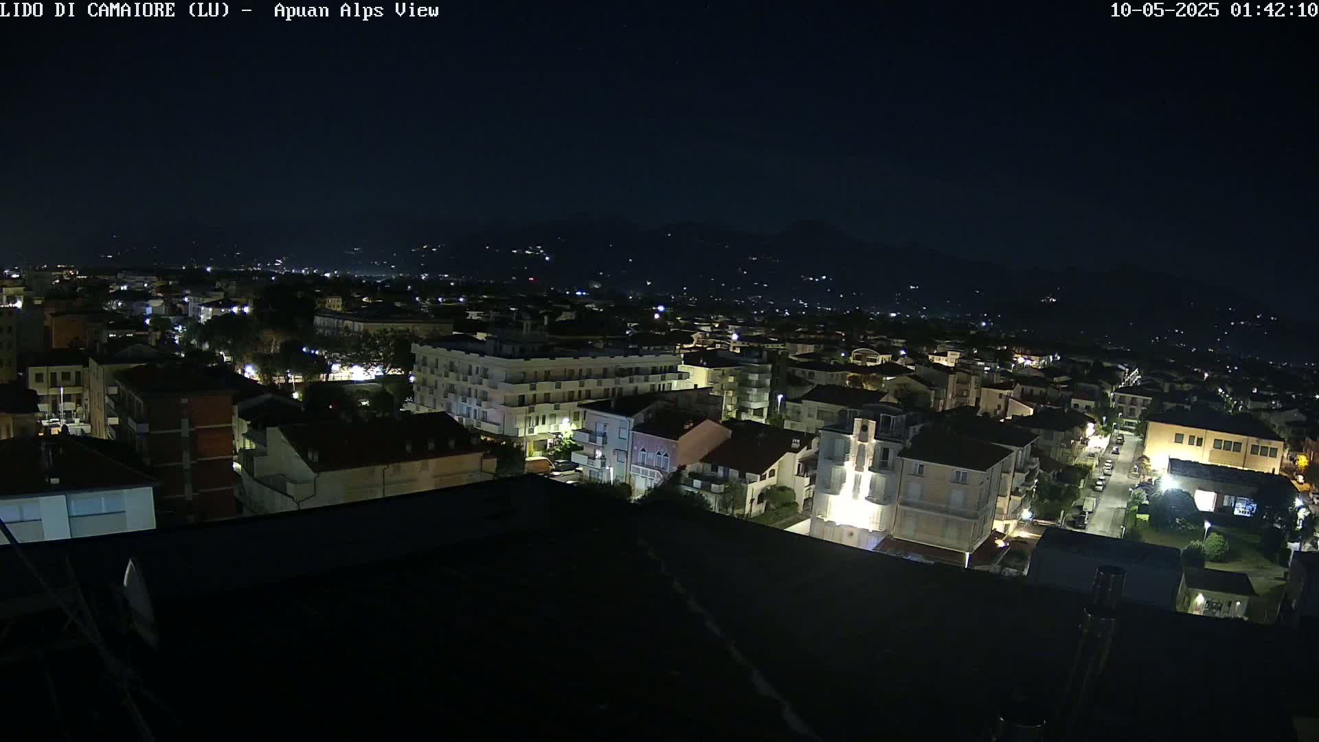 A nighttime, aerial view of a town shows many buildings with illuminated windows, extending to distant, dimly lit hills under a clear, dark sky.