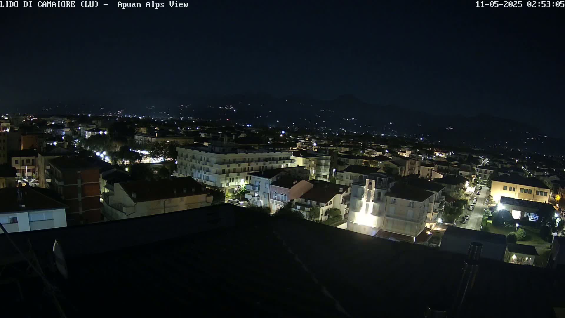A nighttime aerial view of a town illuminated by streetlights and building lights, with dark mountains visible in the distance under a clear night sky.