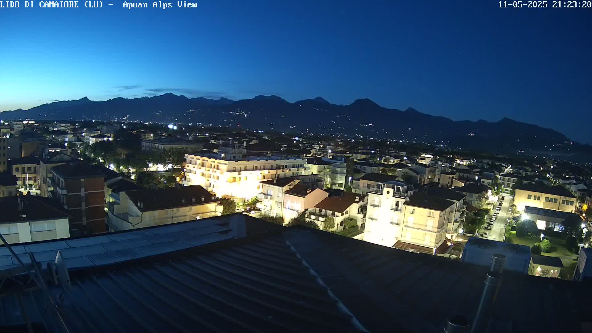 A nighttime view of a town illuminated by streetlights, with a mountain range silhouetted against a dark blue sky.