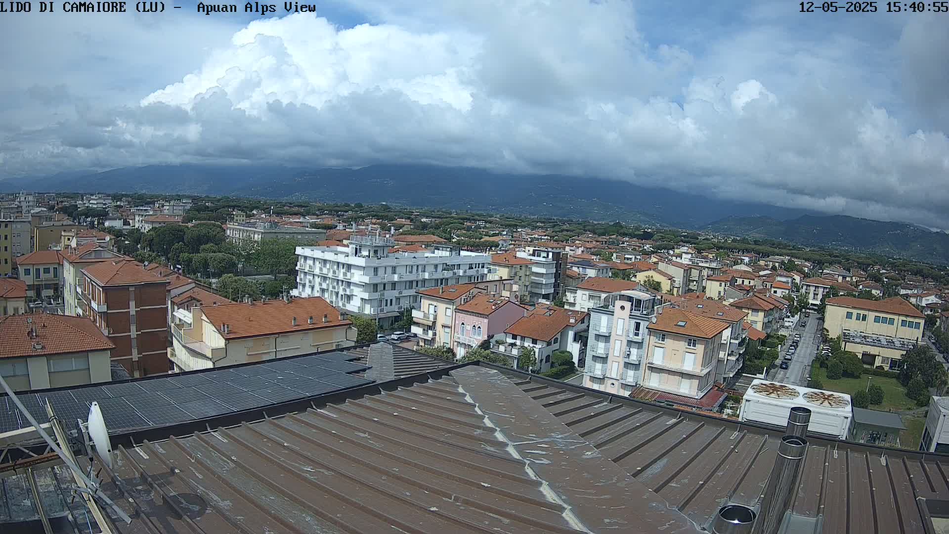 A mostly cloudy sky overlooks a coastal town with red-tiled roofs and distant mountains.