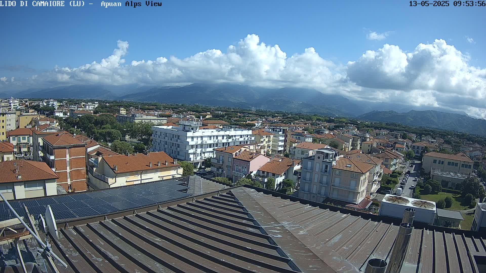 A partly cloudy sky overlooks a town of low-rise buildings nestled against a mountain range.