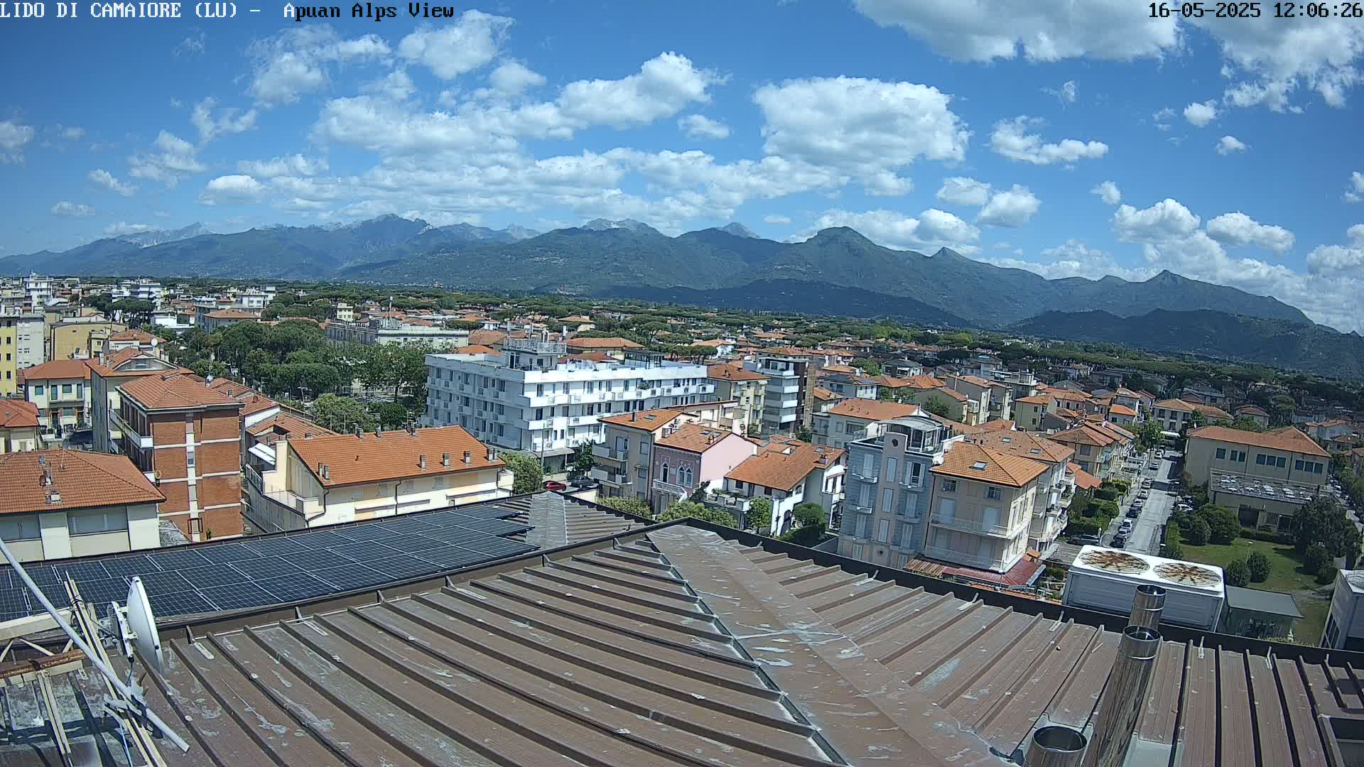 A partly cloudy sky overlooks a town of low-rise buildings with red-tiled roofs, extending to a mountain range in the background.