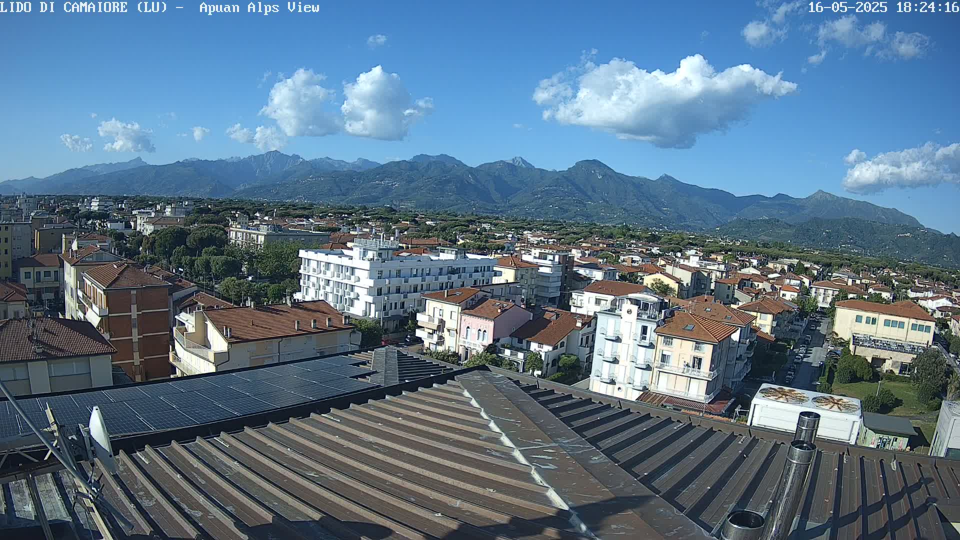 A partly cloudy day reveals a view of a town nestled against a mountain range, seen from a rooftop with solar panels.