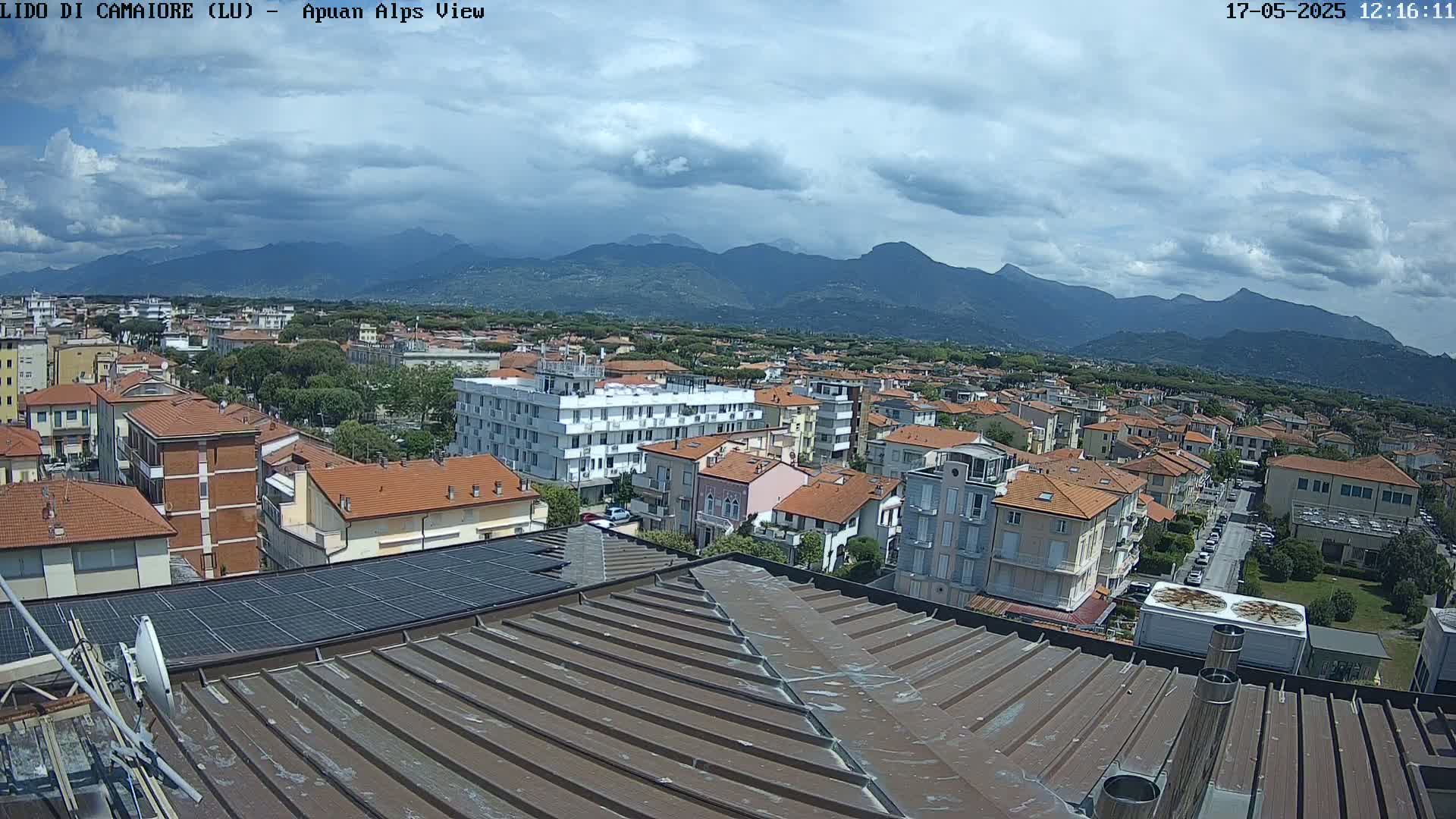 A partly cloudy day reveals a cityscape of low-rise buildings with red tile roofs, stretching towards a mountain range in the distance.