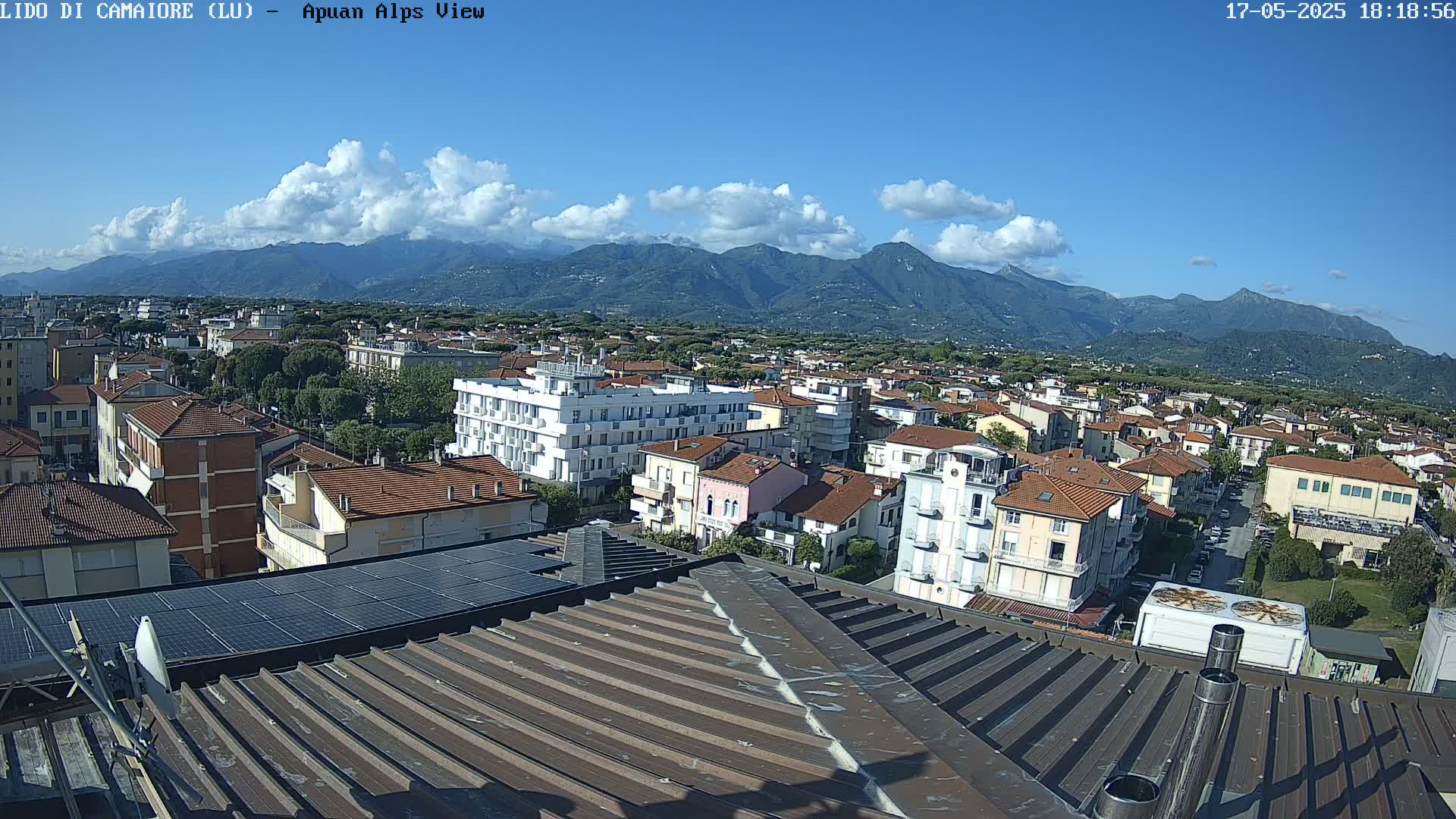 A rooftop view of a town nestled against a mountain range under a mostly sunny sky with some scattered clouds.