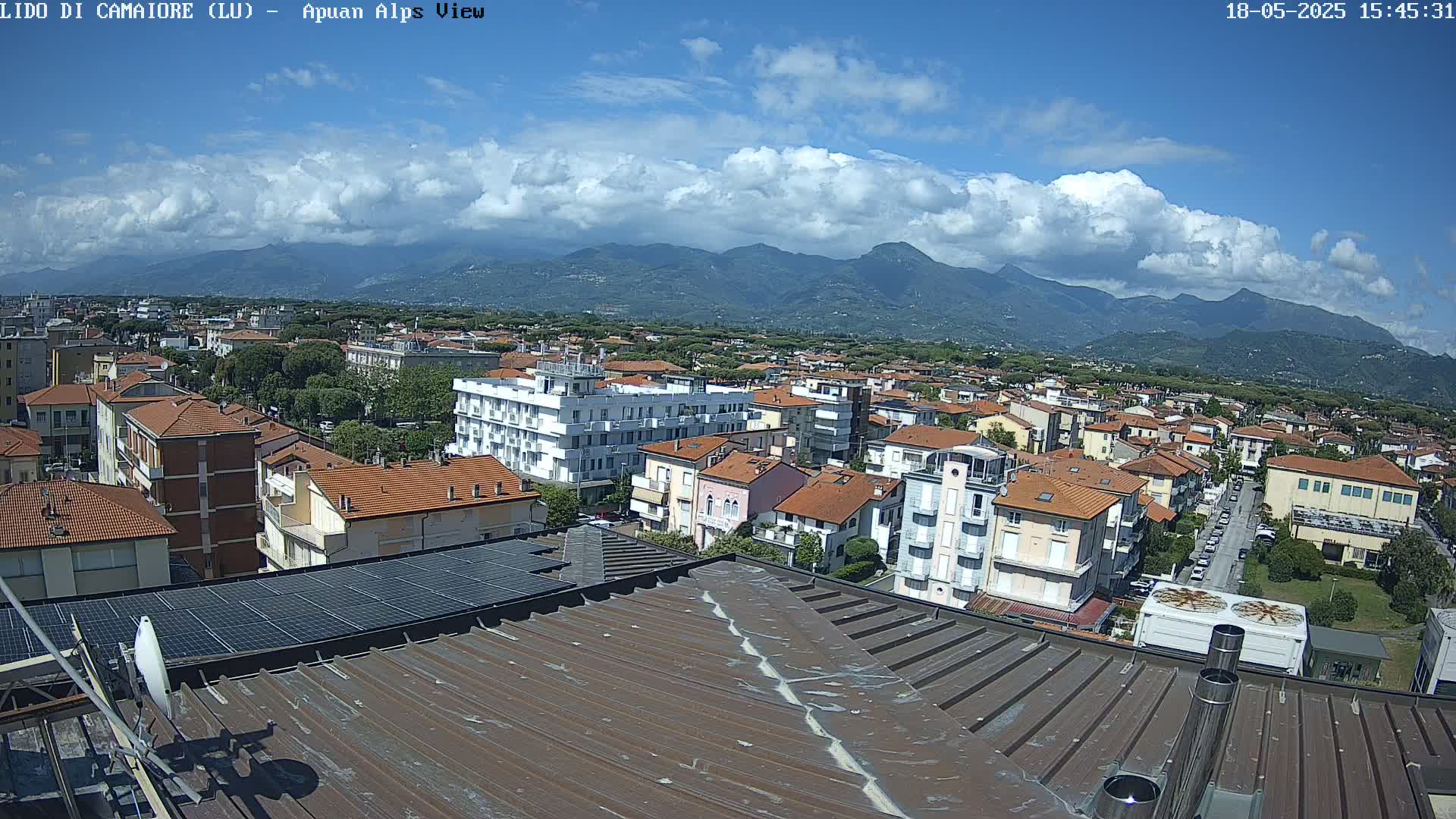 A partly cloudy day reveals a sprawling town nestled against a backdrop of mountains, viewed from a rooftop with solar panels.