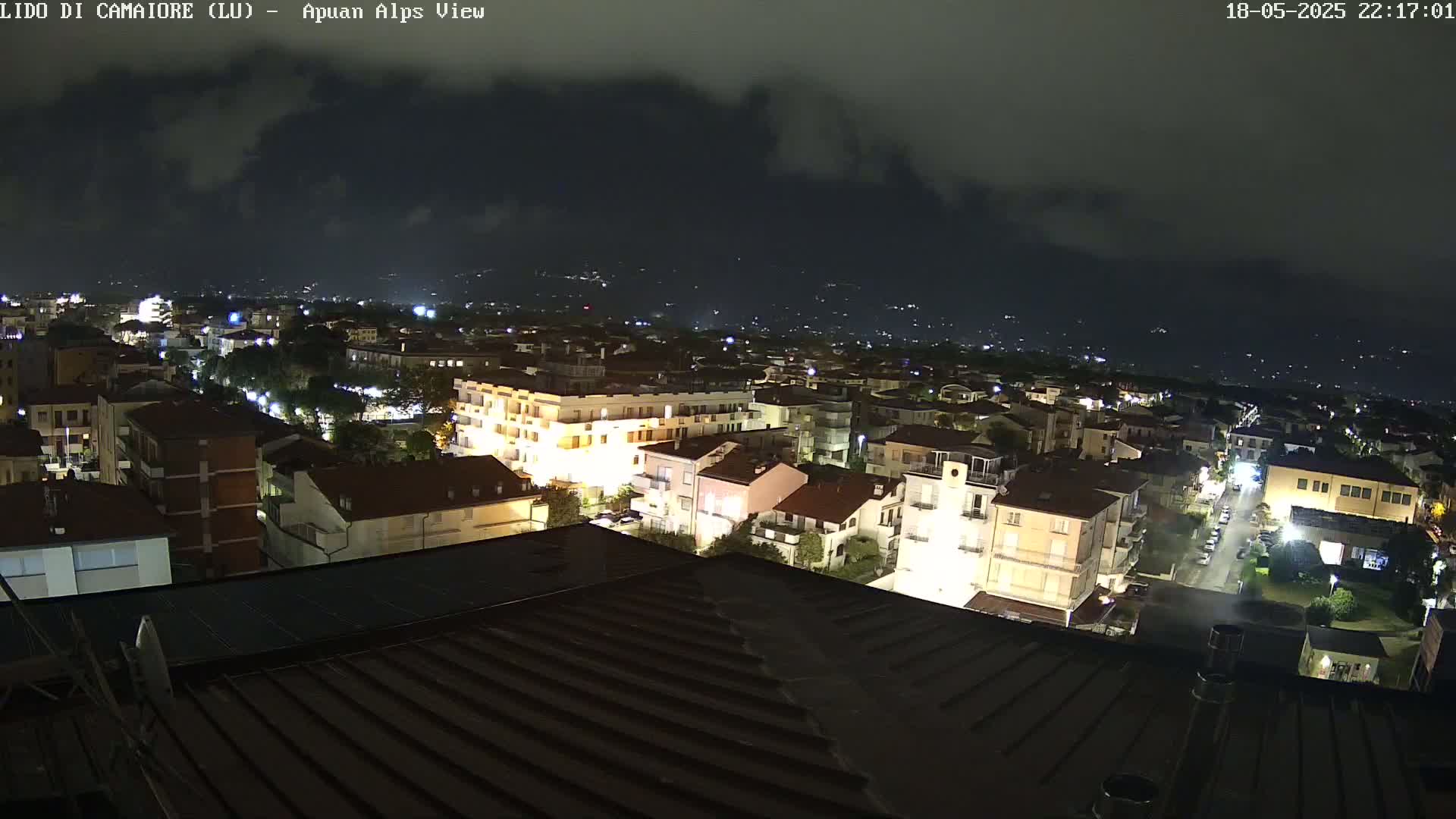 A nighttime, high-angle view of a town illuminated by streetlights and building lights, with a cloudy sky and distant mountains visible in the background.