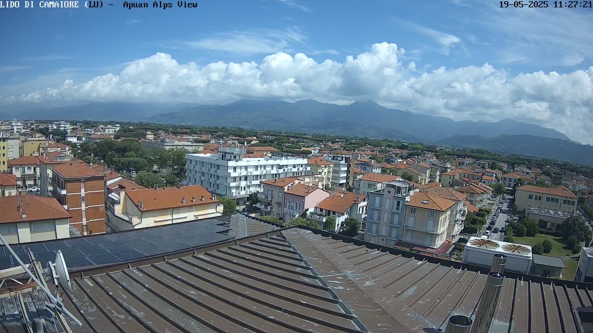 A view from a rooftop shows a town of low-rise buildings under a partly cloudy sky with mountains in the distance.