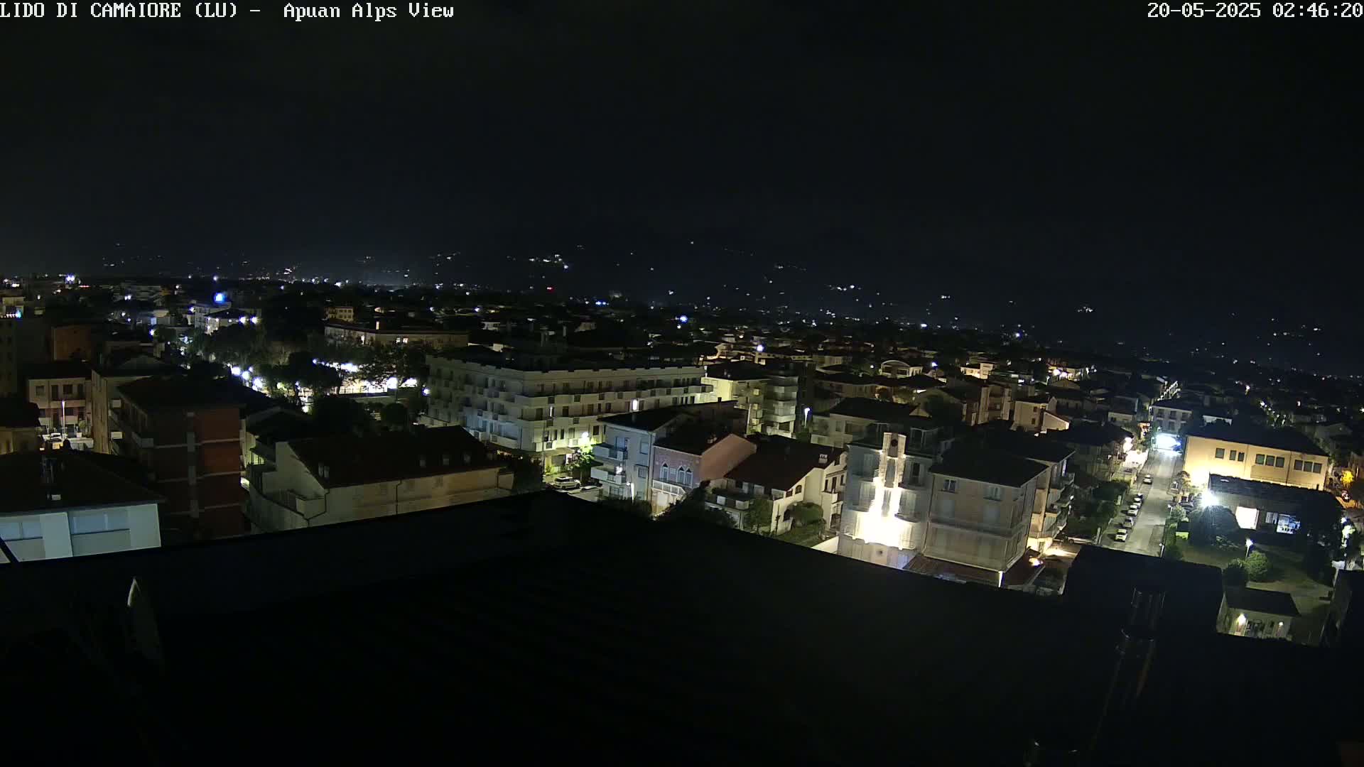 A nighttime aerial view of a town illuminated by streetlights, with a dark sky and distant hills faintly visible in the background.