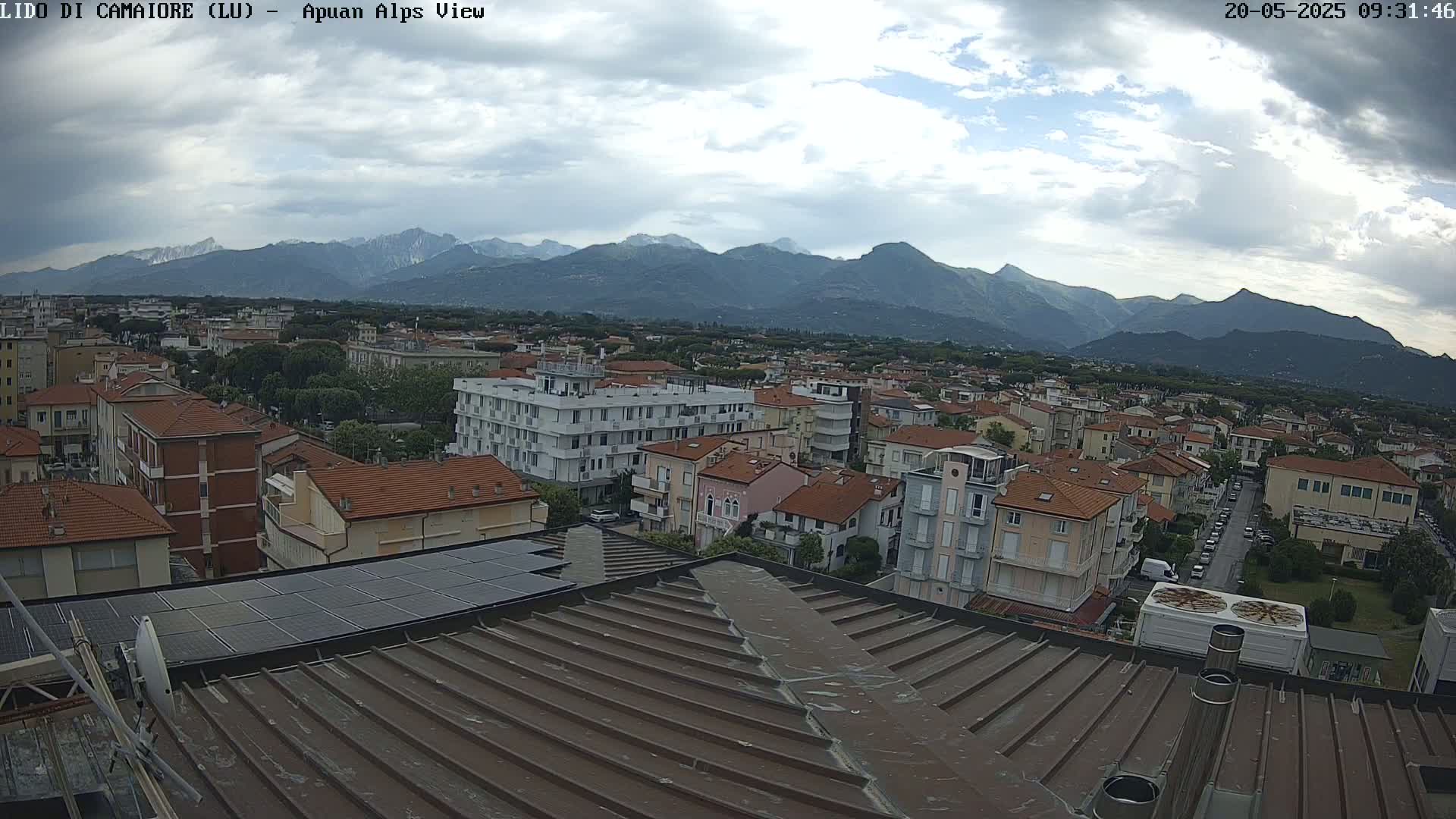 A view from a rooftop shows a town nestled beneath a mountain range under a partly cloudy sky.