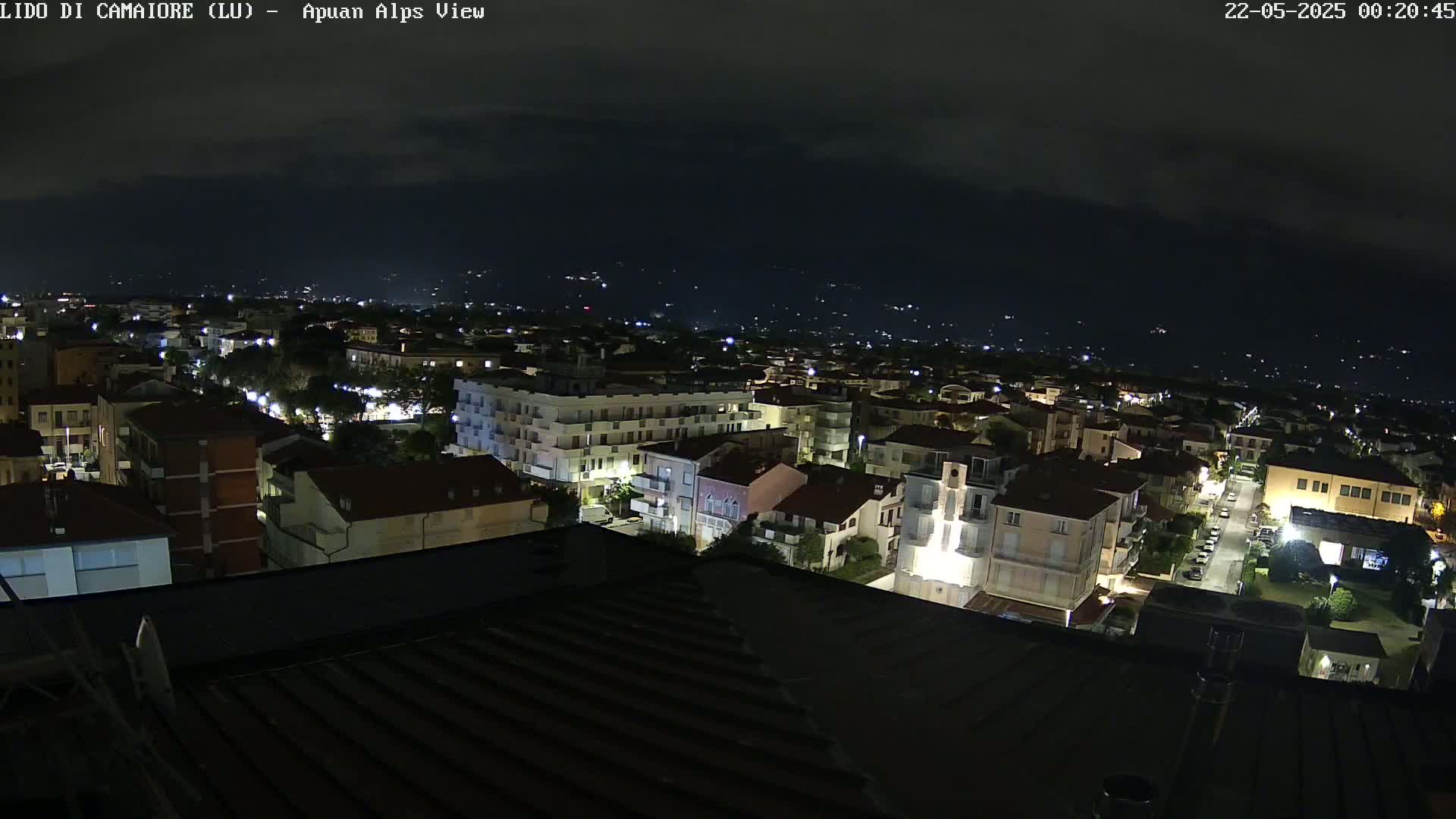 A nighttime aerial view of a town illuminated by streetlights, with a dark mountain range visible in the distance under a mostly clear sky.