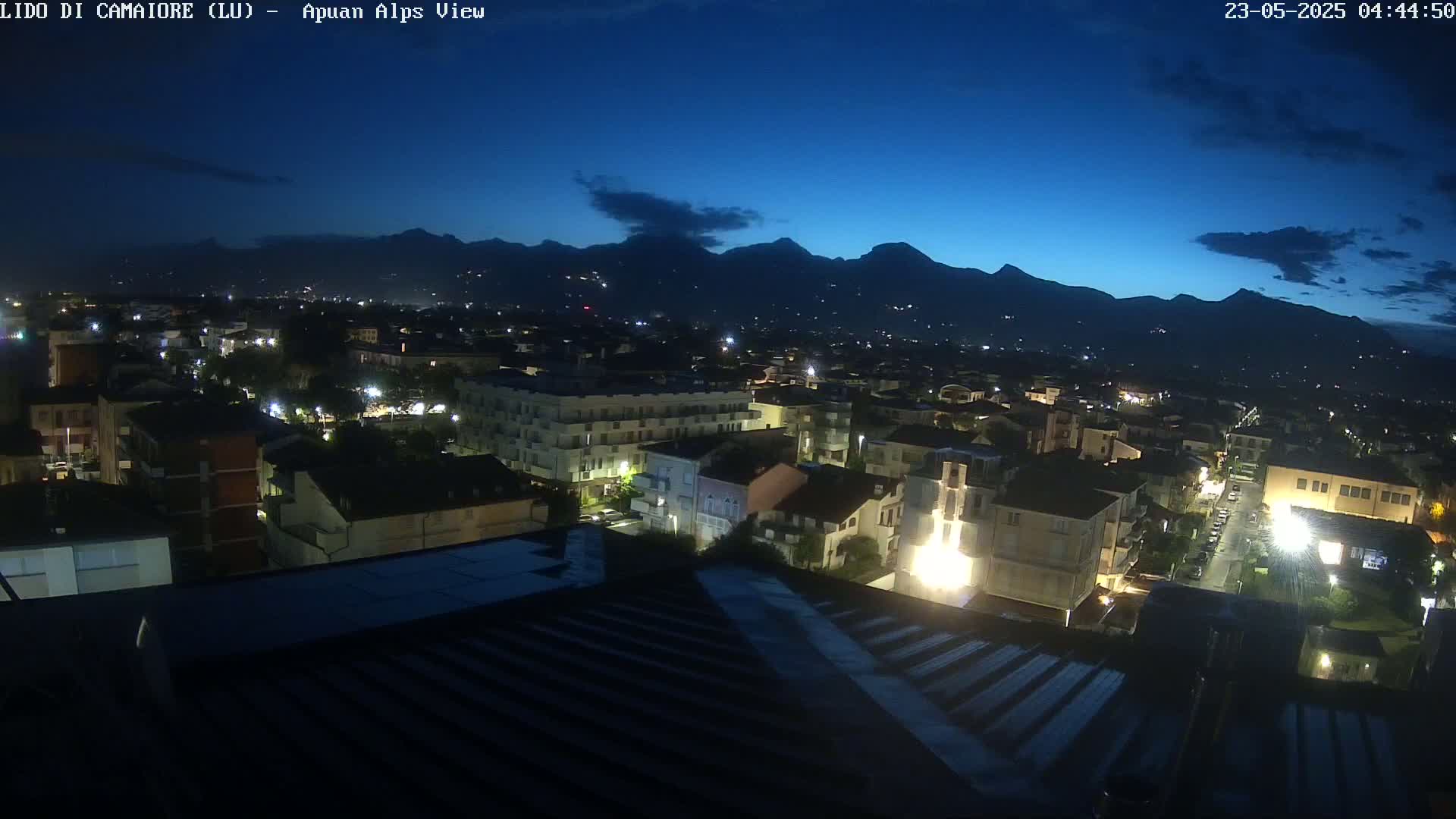 A nighttime view of a town nestled in a valley, illuminated by streetlights, with dark mountains silhouetted against a twilight sky.